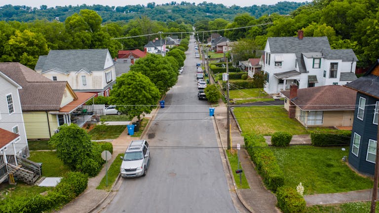 Explore a quiet suburban neighborhood street with houses and lush greenery from an aerial perspective.