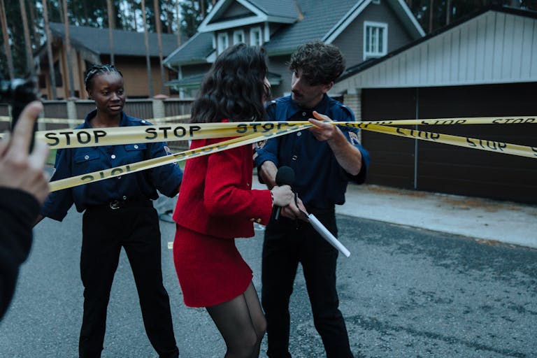 Female journalist interacts with police officers at a taped-off crime scene outdoors.