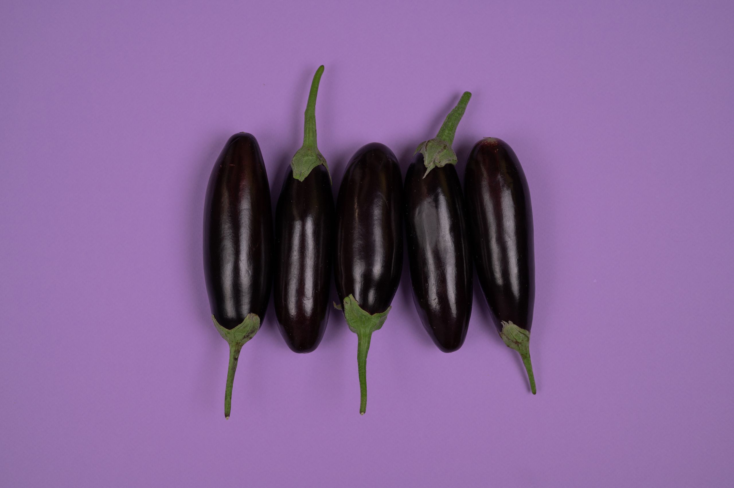Five fresh eggplants aligned on a vibrant purple background, showcasing their deep color and green stems.