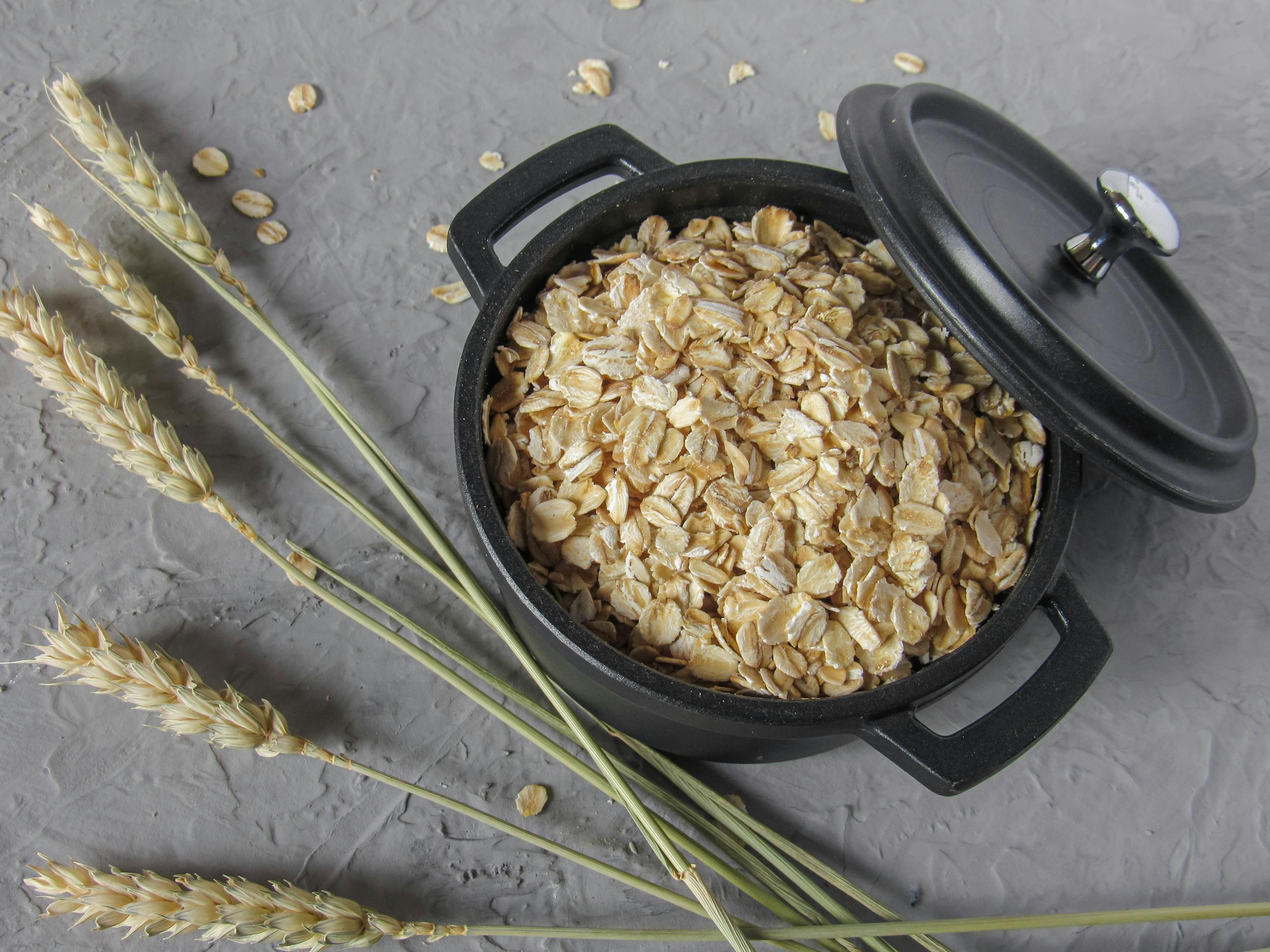 Flat lay of rolled oats in a black pot surrounded by wheat stalks on a textured surface.