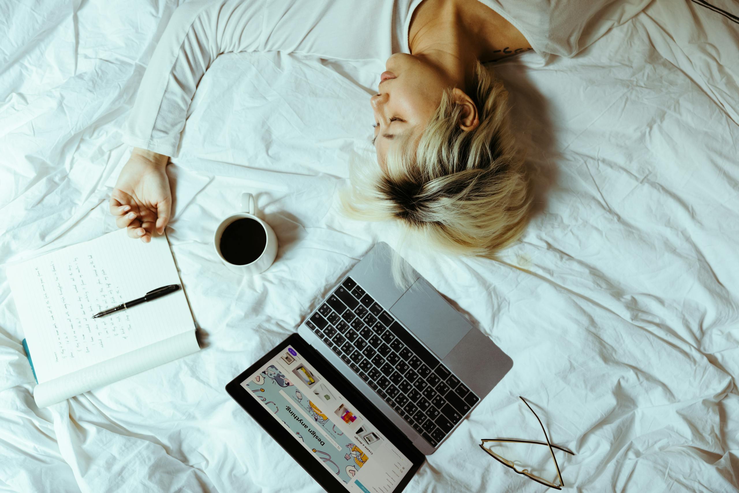 From above of exhausted female student lying with closed eyes on crumpled bed sheet near netbook and open notebook with pen and cup of coffee at home