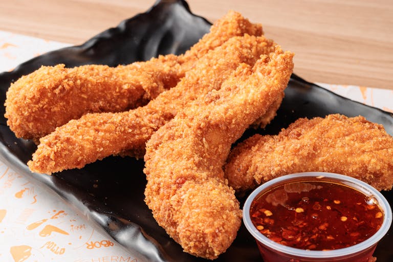Golden brown fried chicken strips served with a spicy chili dip on a black plate.