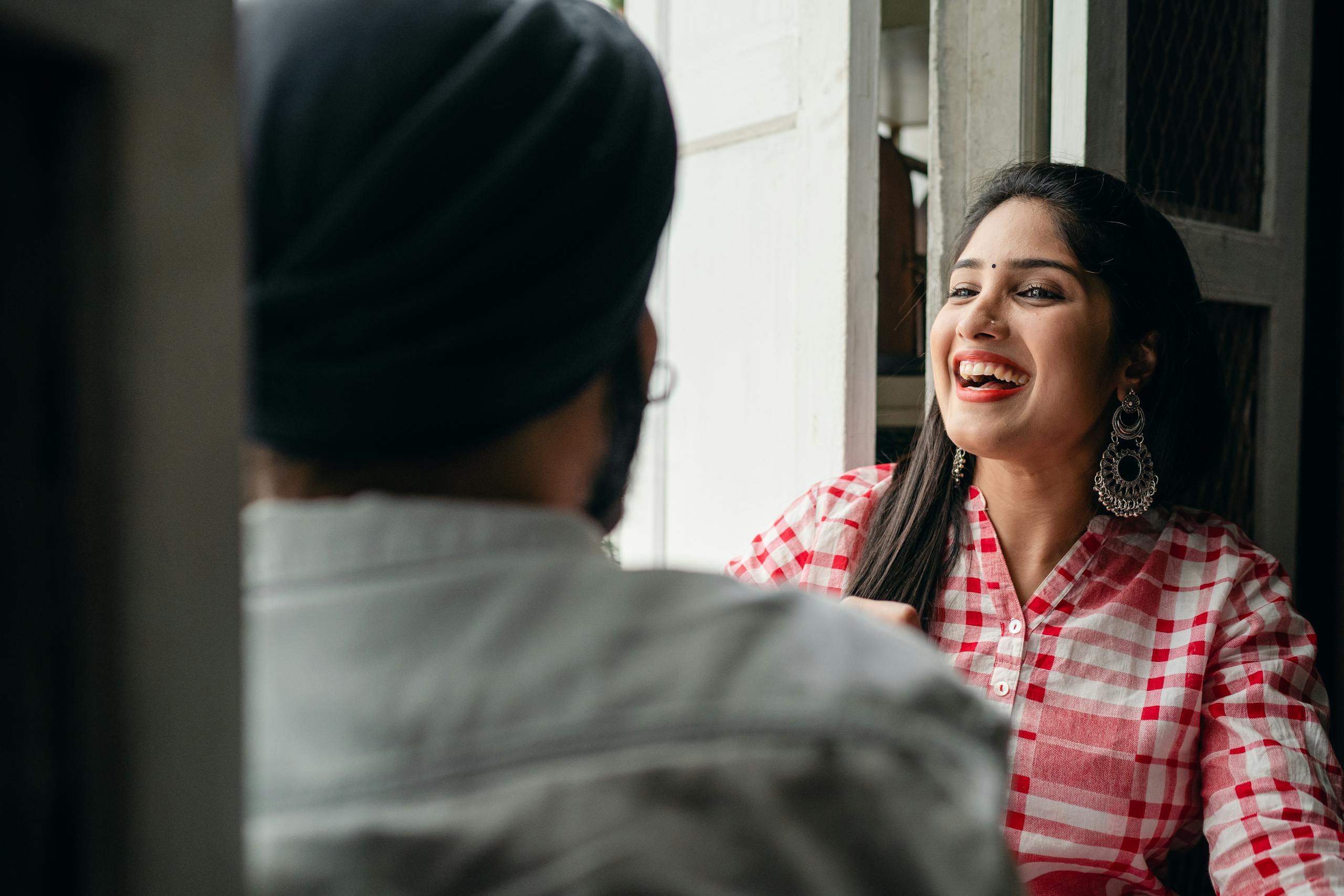Good looking Indian female in checkered shirt with massive earrings laughing happily while listening to funny stories told by unrecognizable man in turban