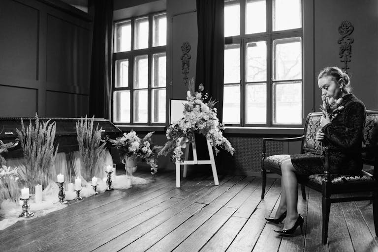 Grayscale funeral scene showing a woman in mourning by a coffin and floral arrangements.