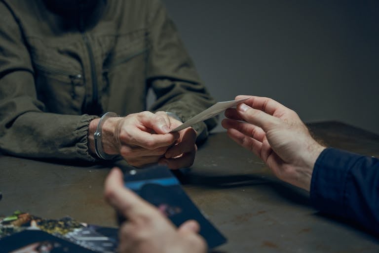 Hands exchanging evidence during an interrogation scene in a dimly lit room.