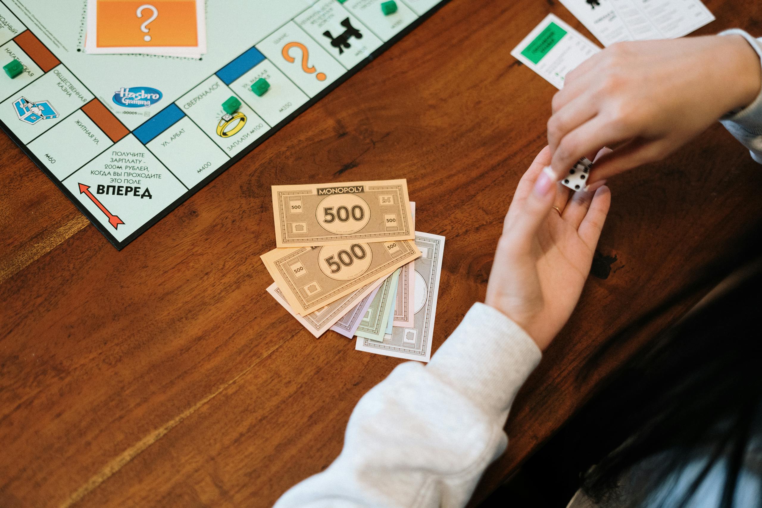 Hands are holding dice near Monopoly board with fake money on table.