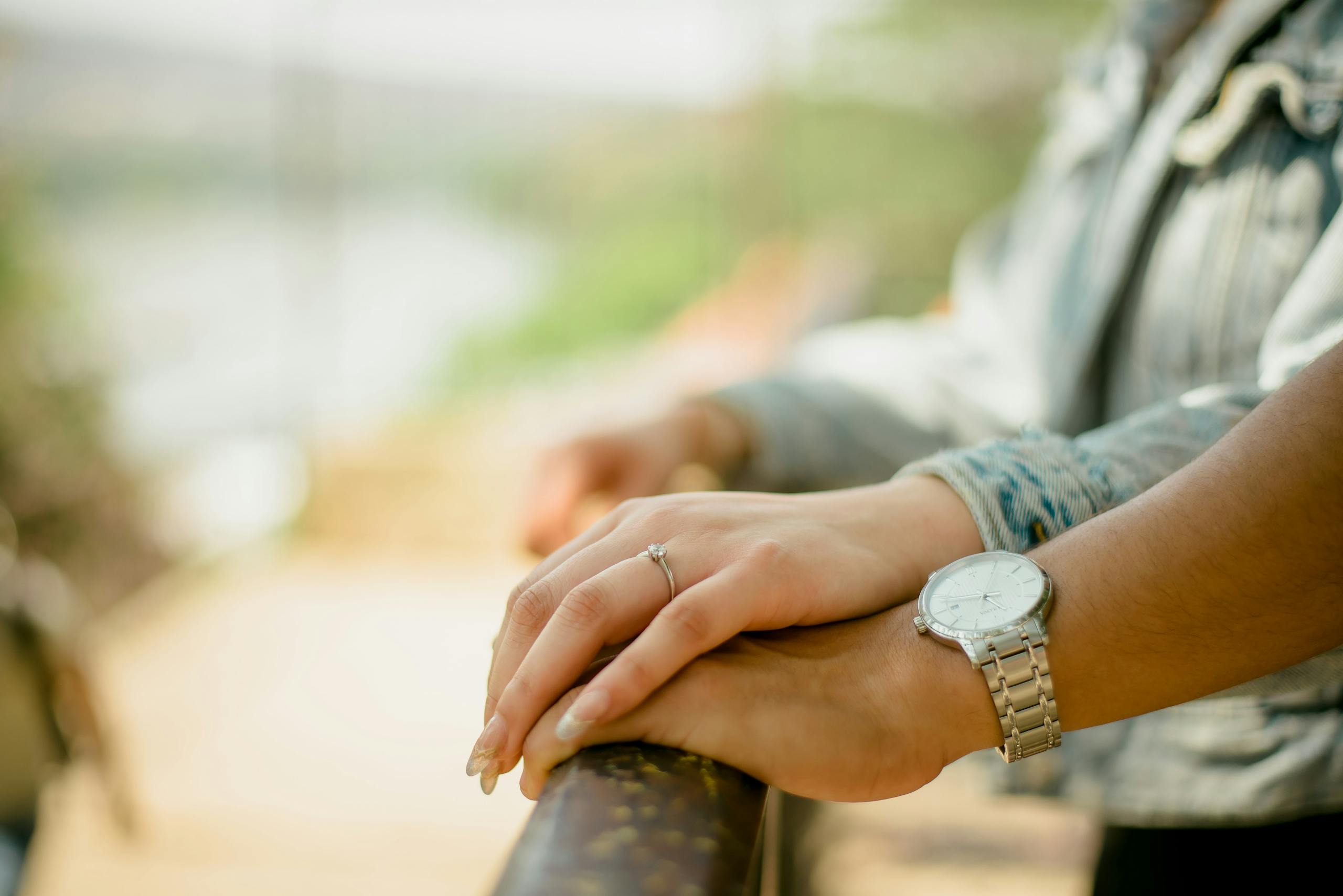 Hands of a couple resting on a railing, showcasing an engagement ring and a wristwatch.