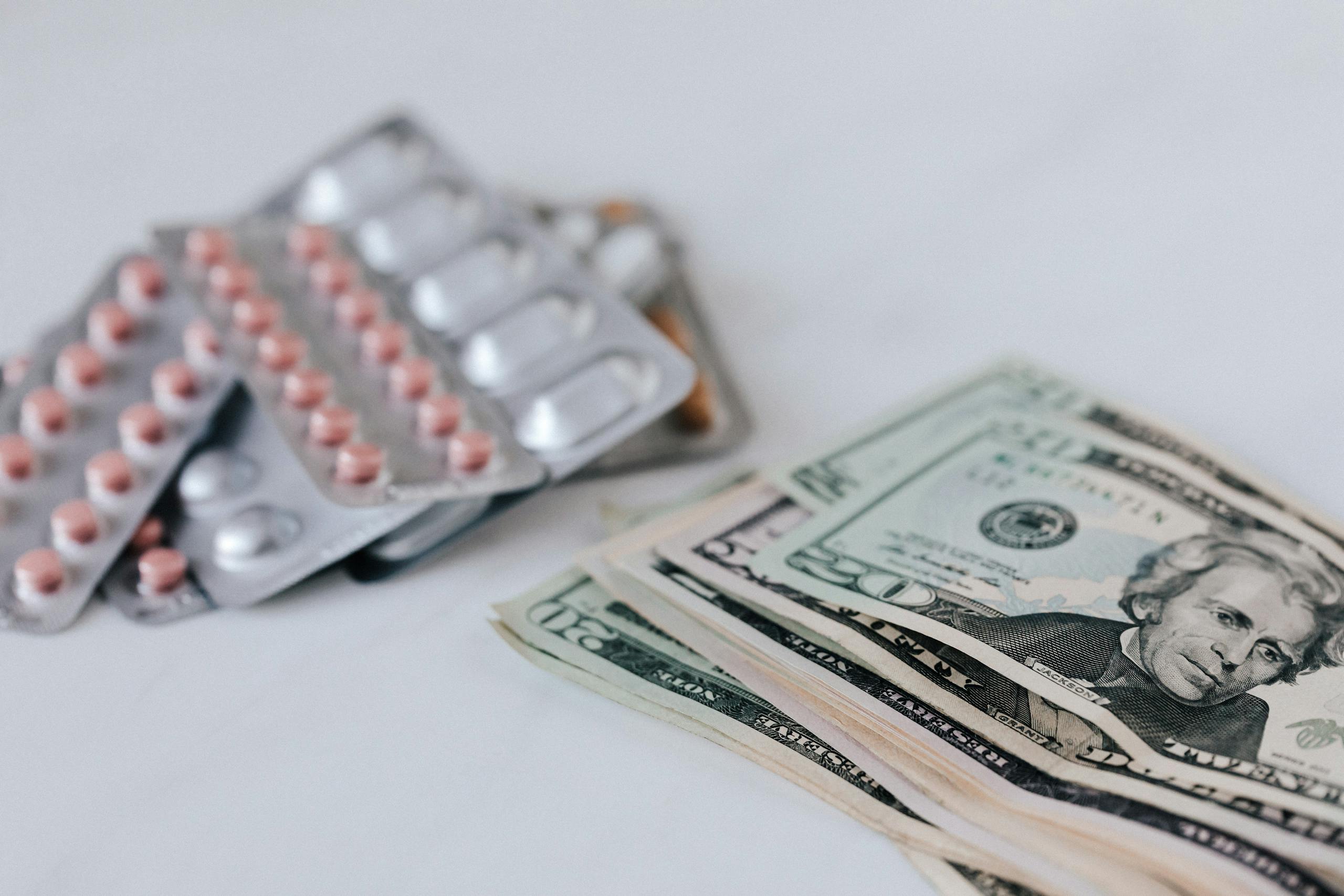 Heap of similar American dollars in cash placed near blisters of expensive drugs on white table