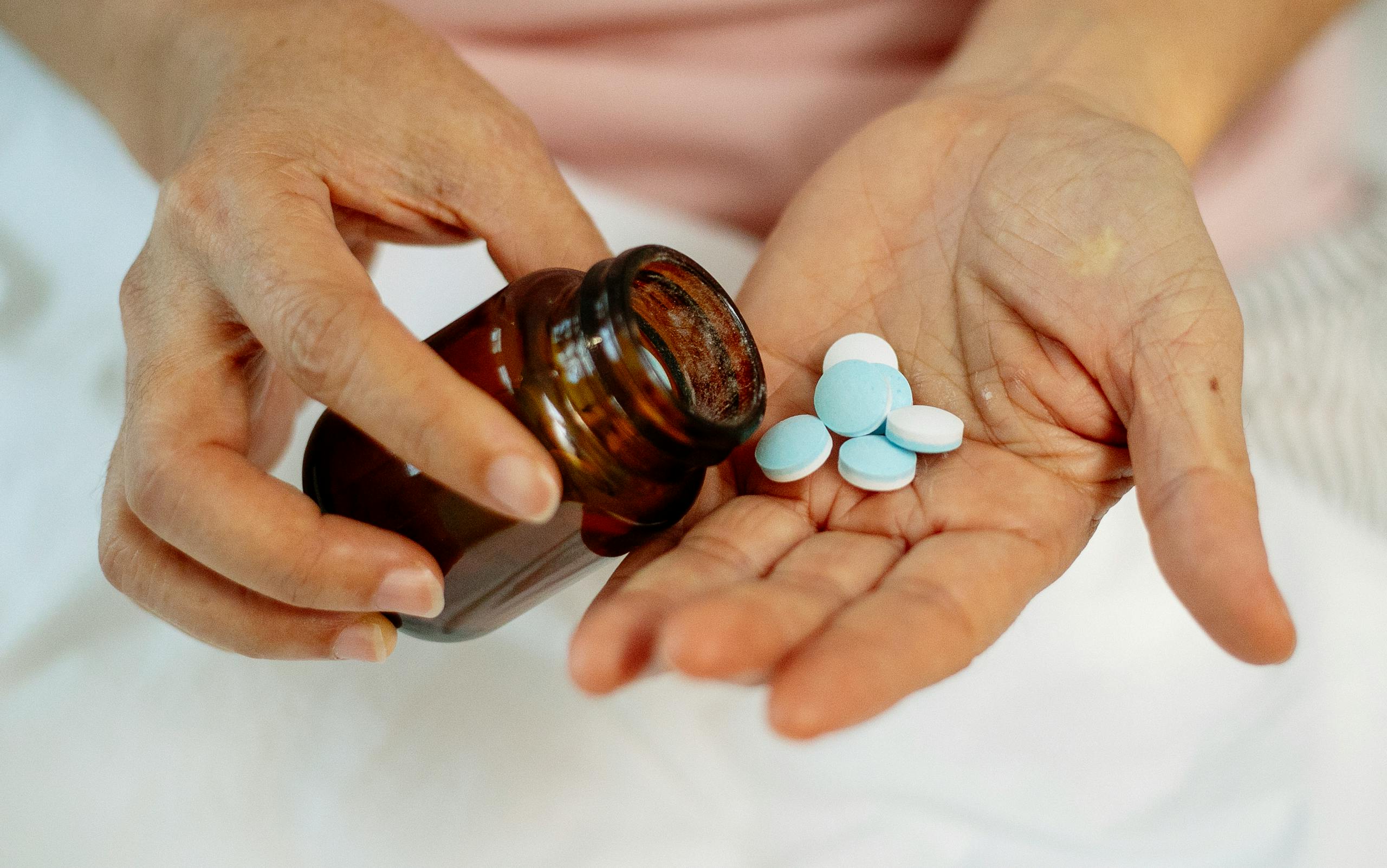 High angle of crop faceless woman under blanket with white and blue pills in hand and glass bottle in arm in bright room