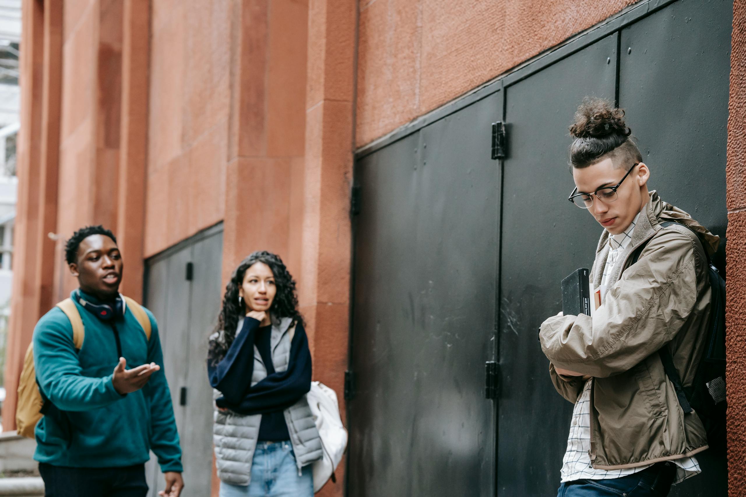 Low angle of aggressive young diverse groupmates bullying shy upset male leaning on building and looking down