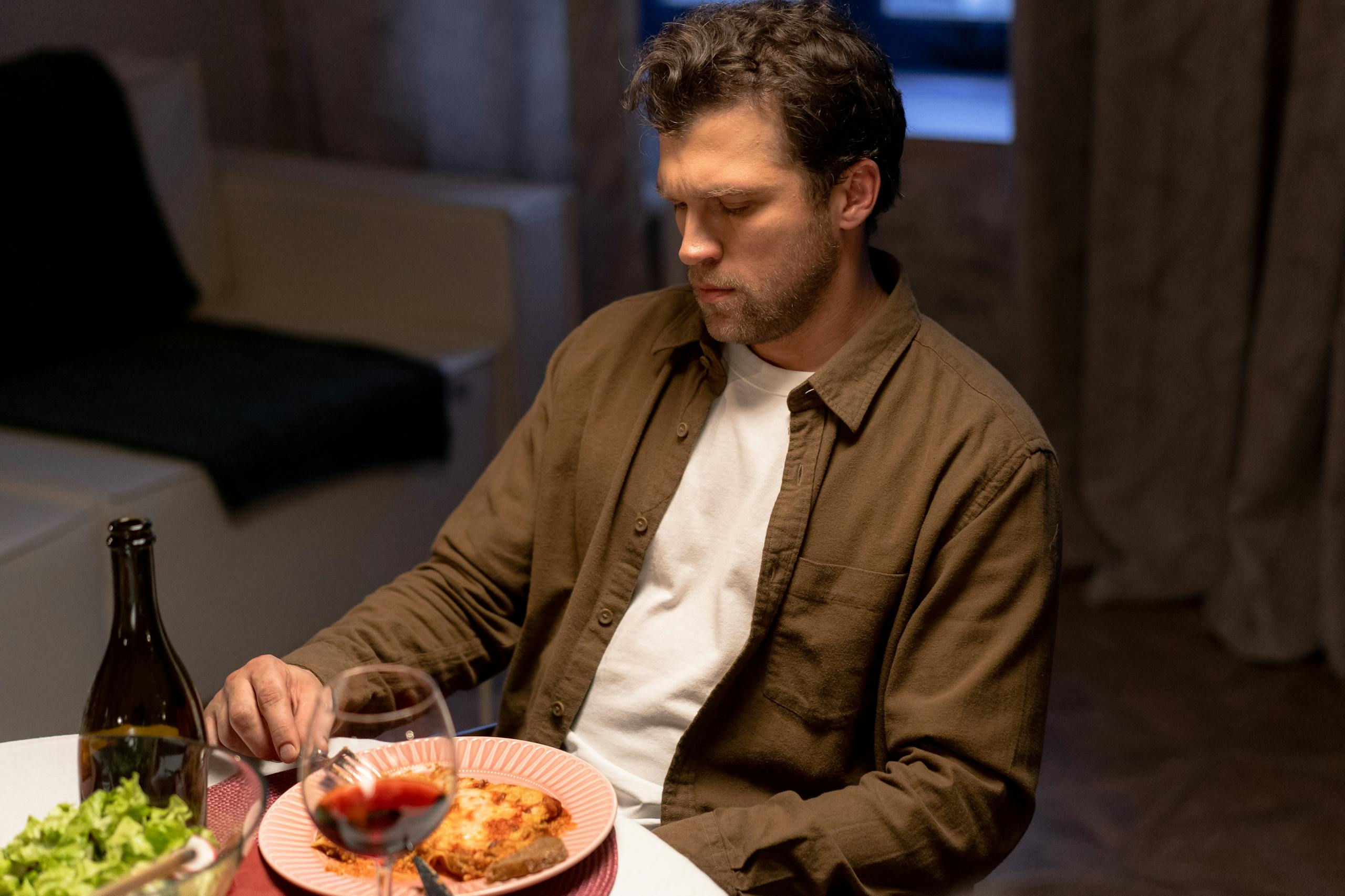 Man seated at dinner table with wine, lasagna, and salad, appearing thoughtful.