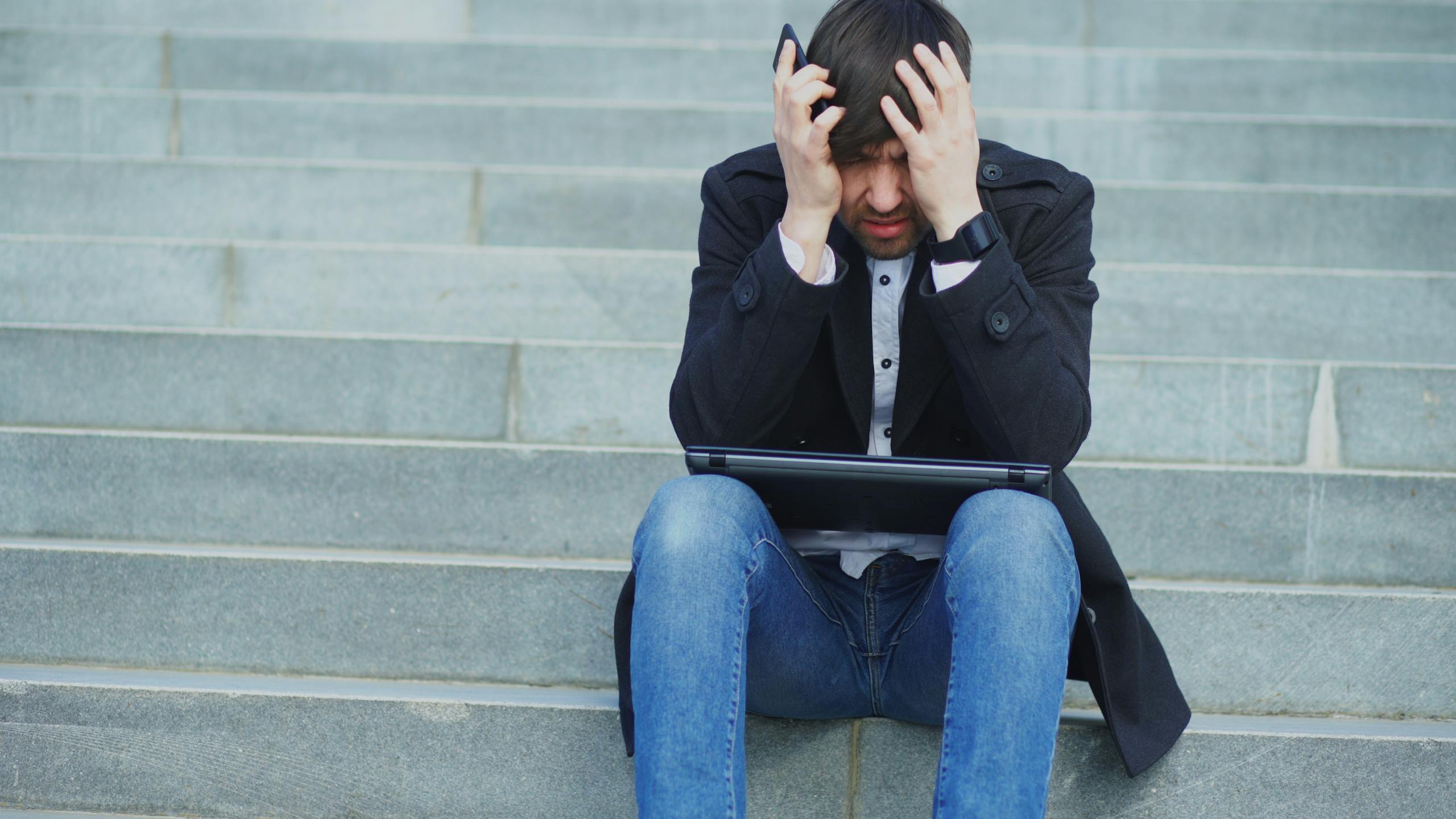Man sitting on stairs with laptop, looking stressed and frustrated.