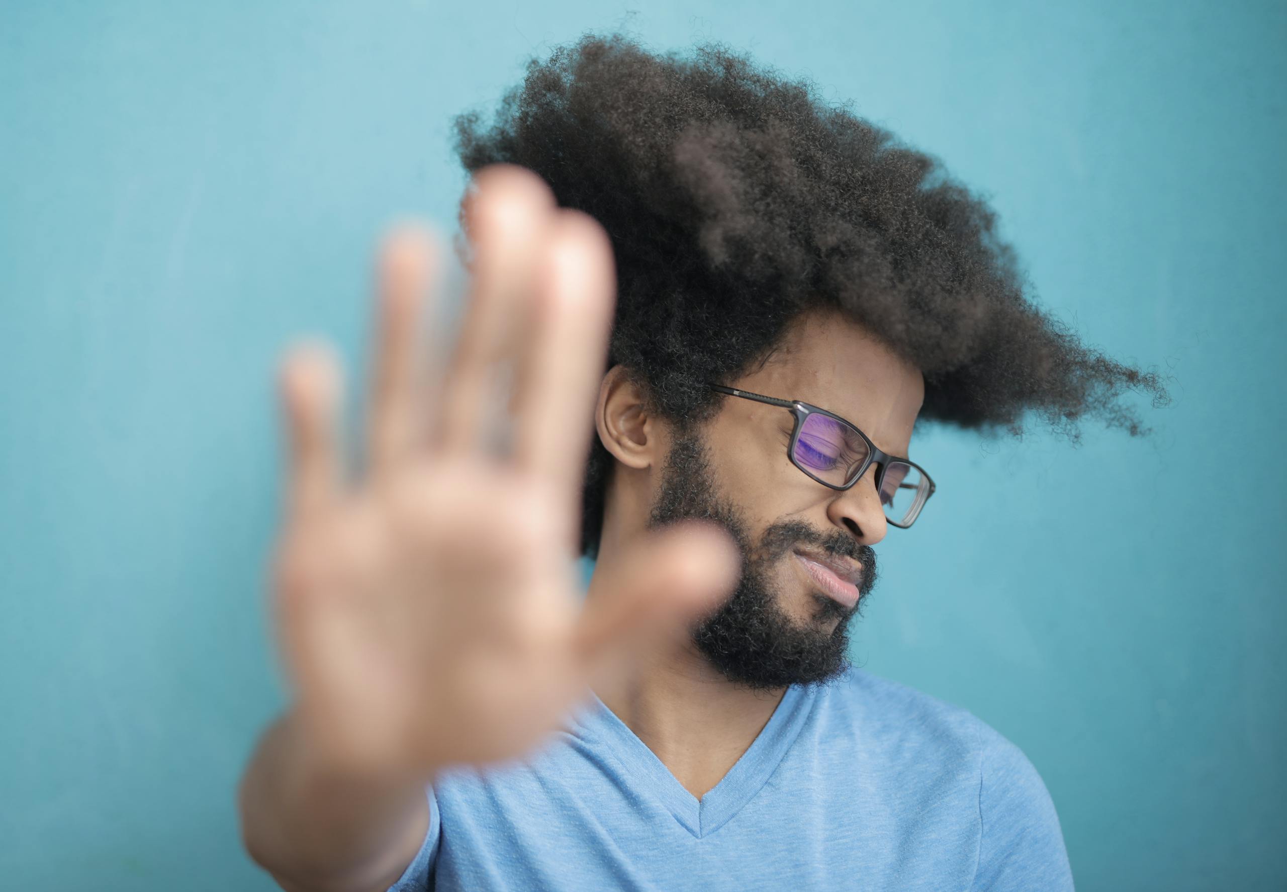 Man with afro hair and glasses making a stop gesture against blue background.
