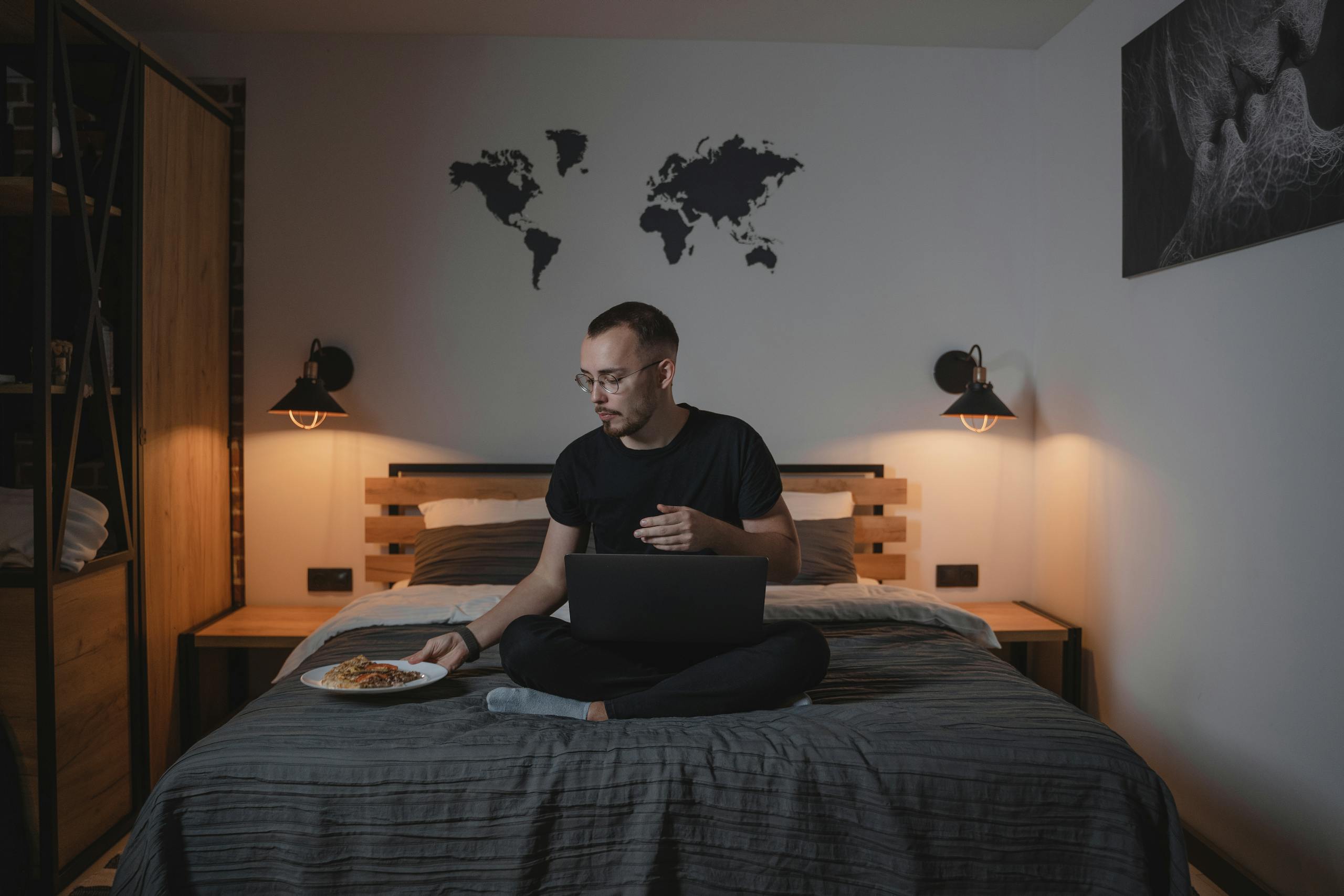 Man working remotely on a bed with a laptop and pizza in a stylish, modern bedroom.