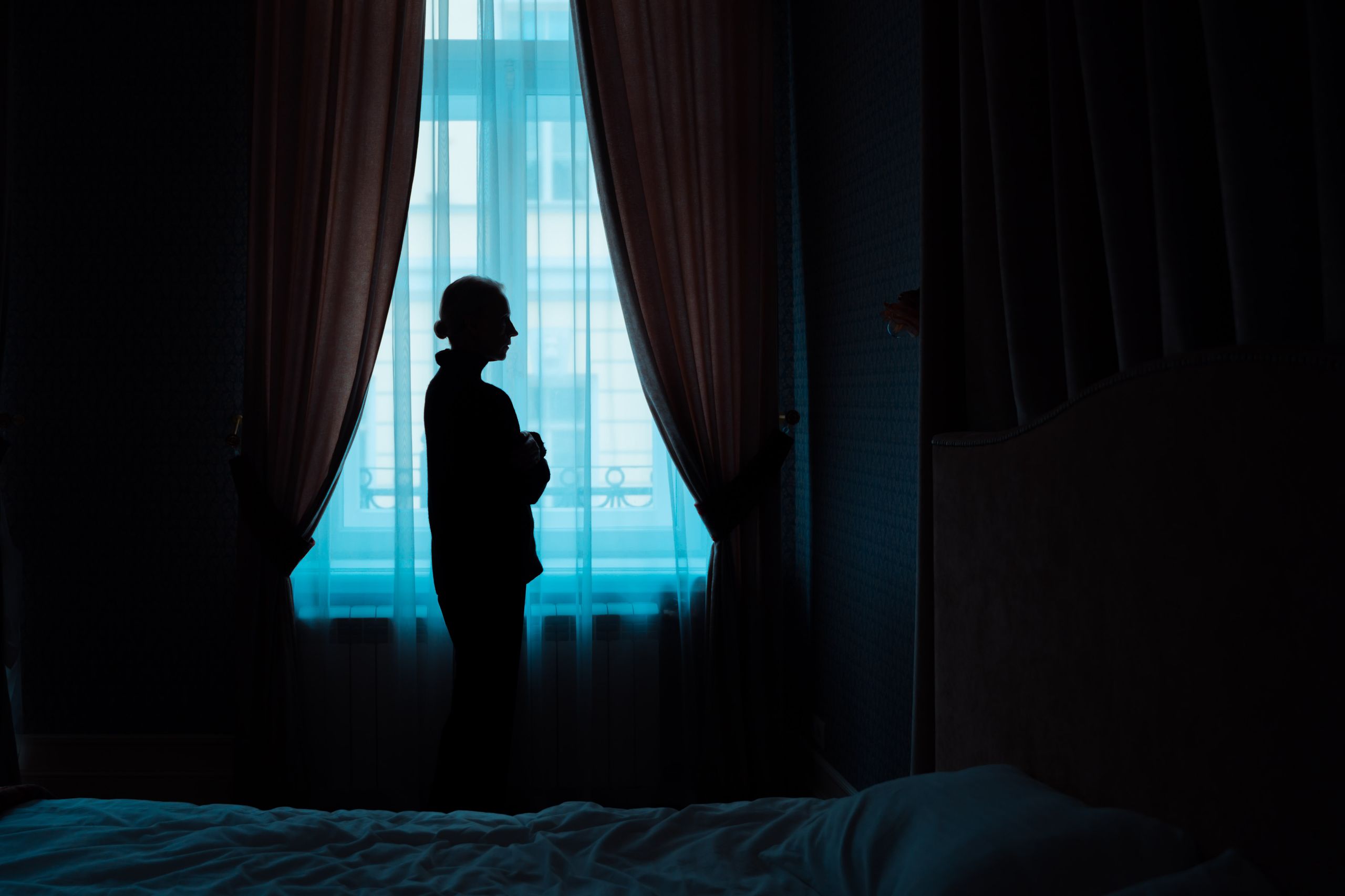 Moody silhouette of a woman standing alone by a window with curtains in a dimly lit room.