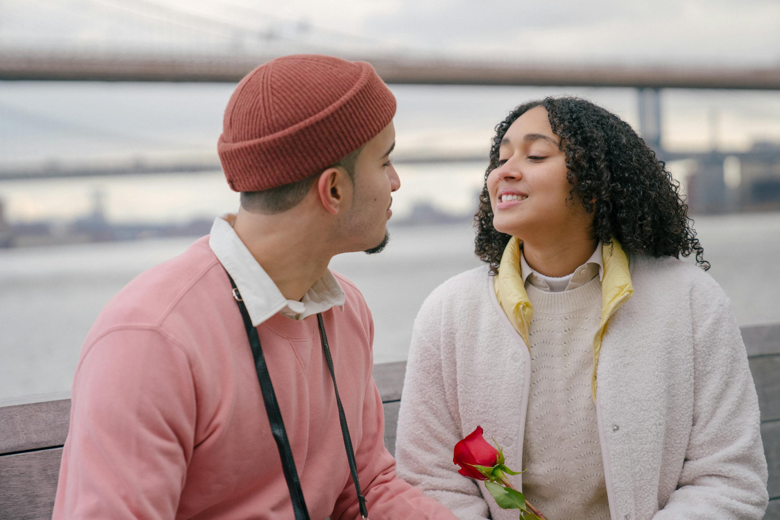 Optimistic Hispanic girlfriend with red rose and boyfriend looking at each other while sitting on bench on embankment during date against blurred background