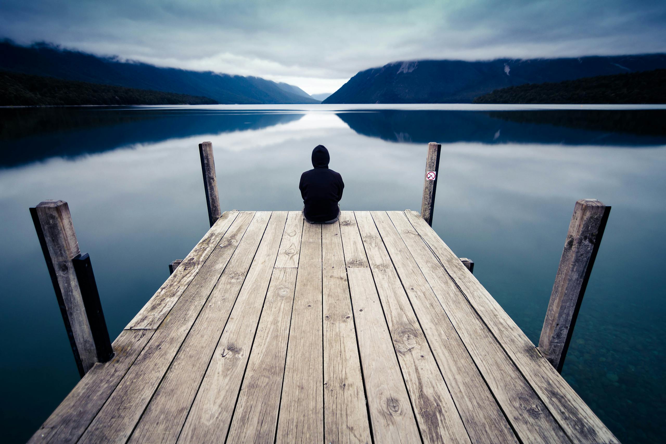 Person sitting in solitude on a wooden dock in New Zealand's serene landscape, surrounded by calm waters.