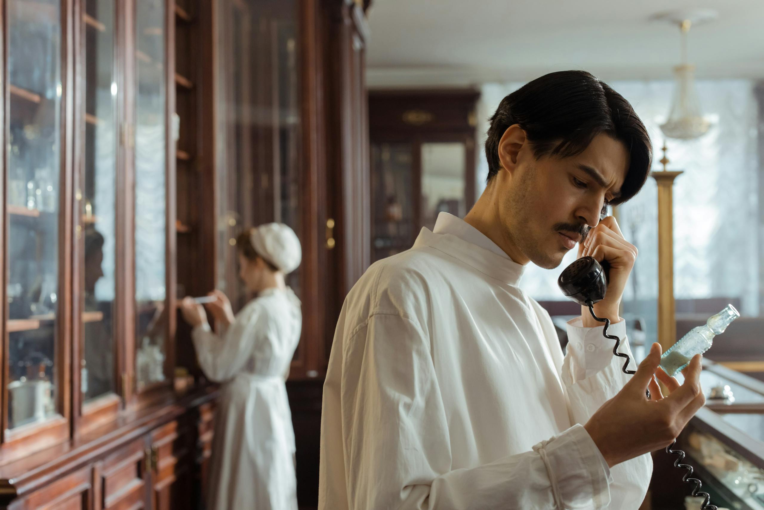 Pharmacist examining a flask while speaking on a vintage telephone in a classic pharmacy setting.