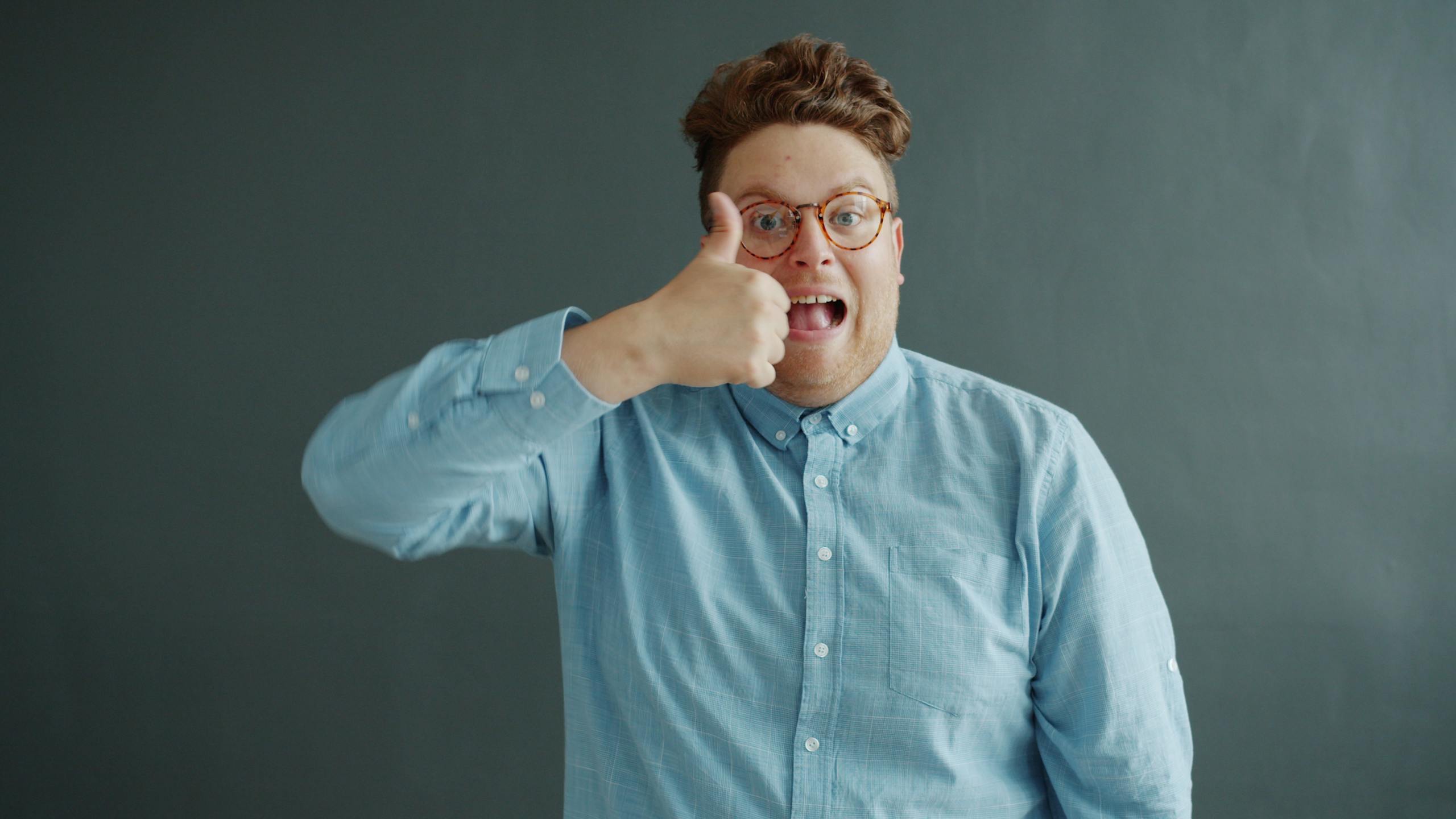 Portrait of a cheerful man in glasses giving a thumbs up against a plain background.