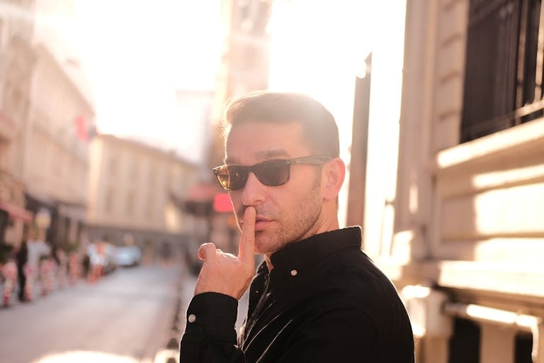Portrait of a man wearing sunglasses making a silence gesture outdoors in bright sunlight.