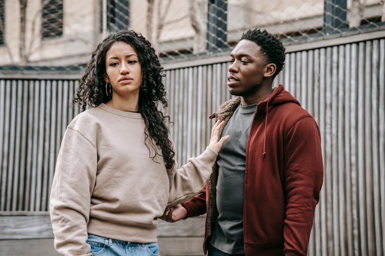 A sad ethnic girlfriend with curly hair rejecting annoyed African American boyfriend while arguing on street near wooden fence during breakup