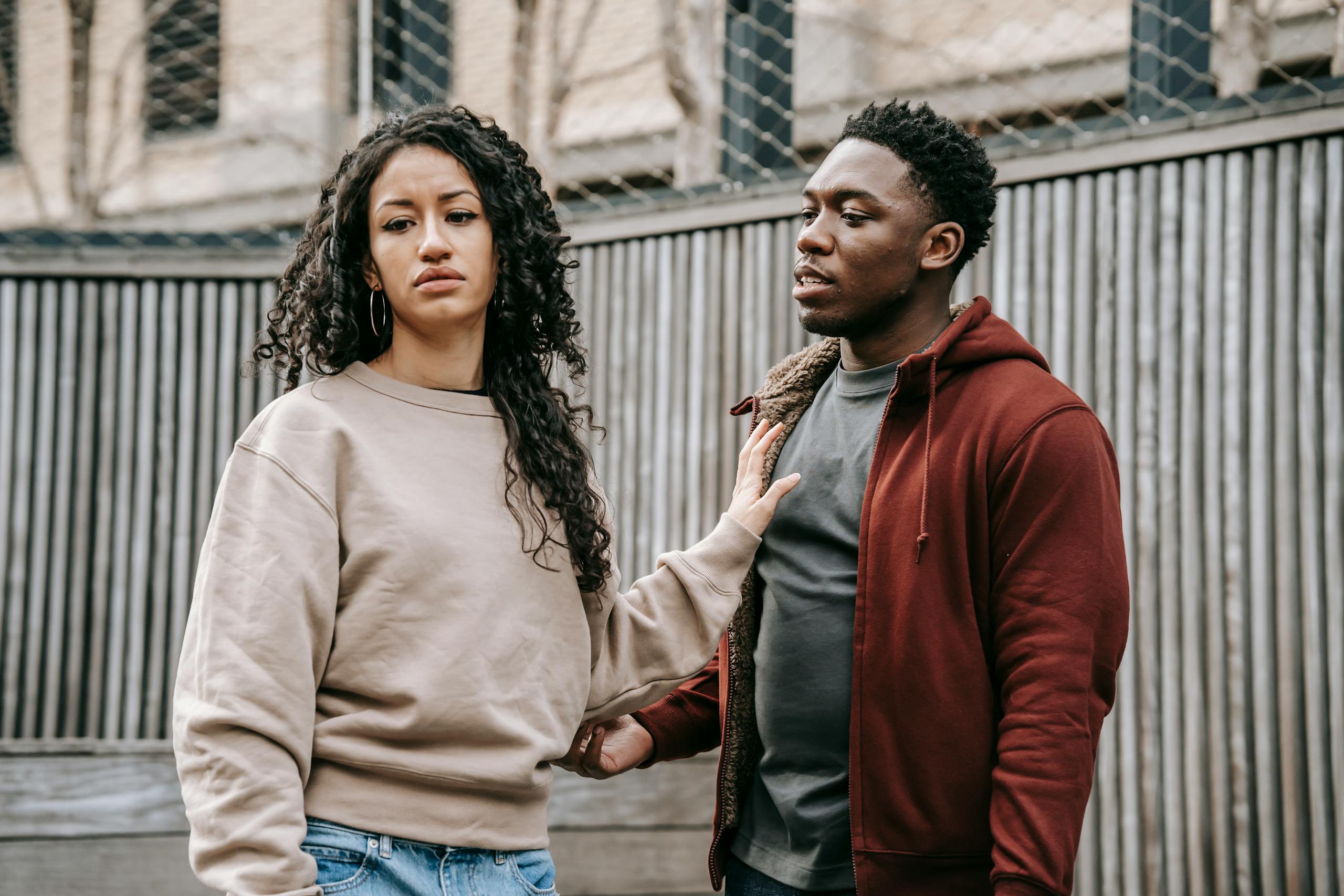 A sad ethnic girlfriend with curly hair rejecting annoyed African American boyfriend while arguing on street near wooden fence during breakup