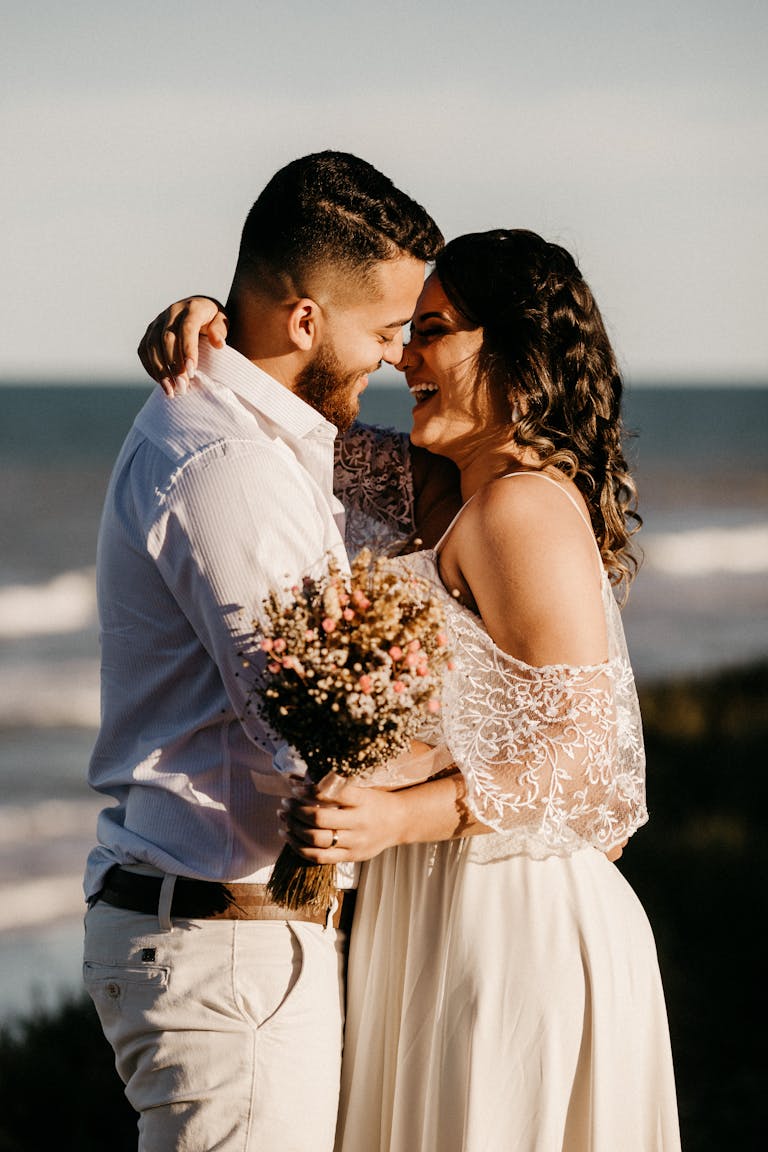 Side view of cheerful young ethnic newlywed couple embracing and laughing while standing on shore near waving ocean during wedding celebration