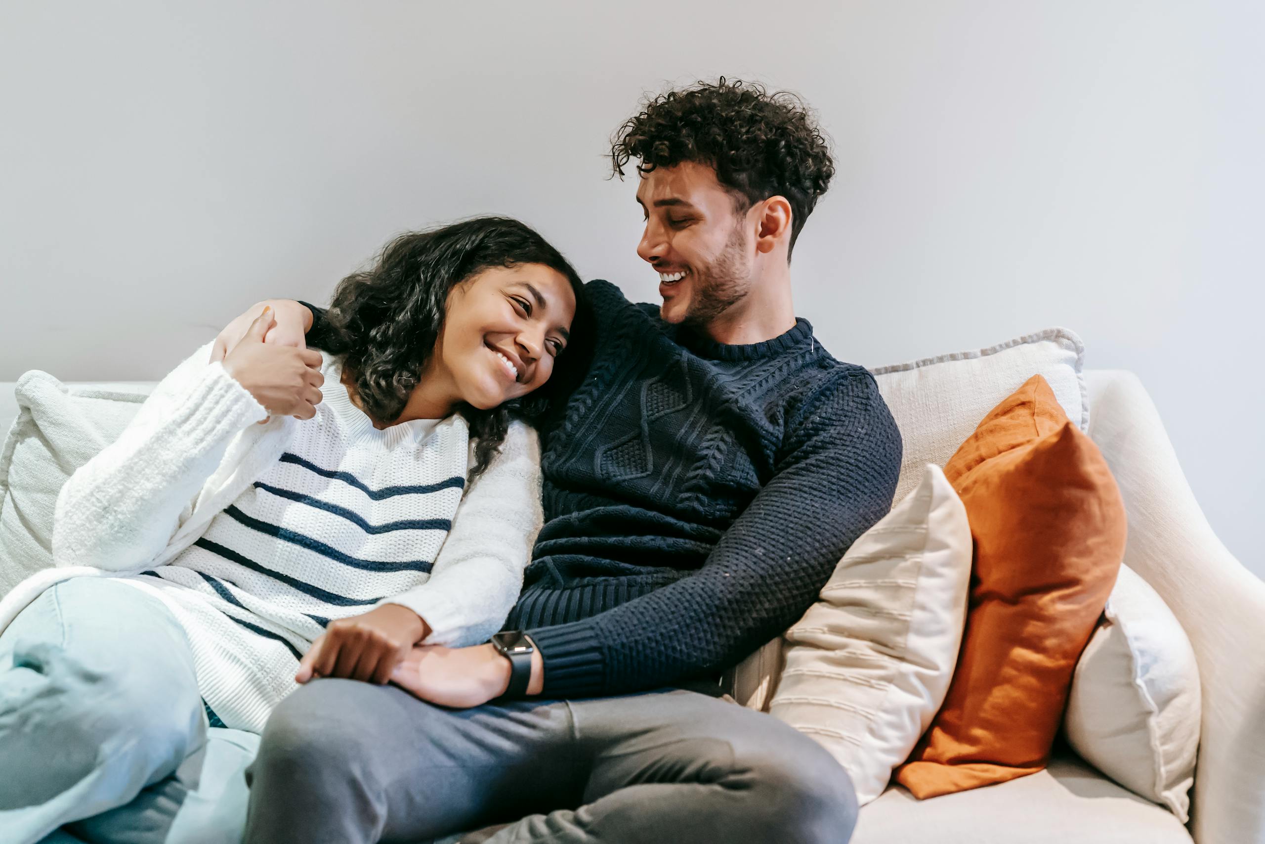 Smiling African American girlfriend and positive ethnic boyfriend wearing casual clothes embracing on comfortable sofa in living room during free time