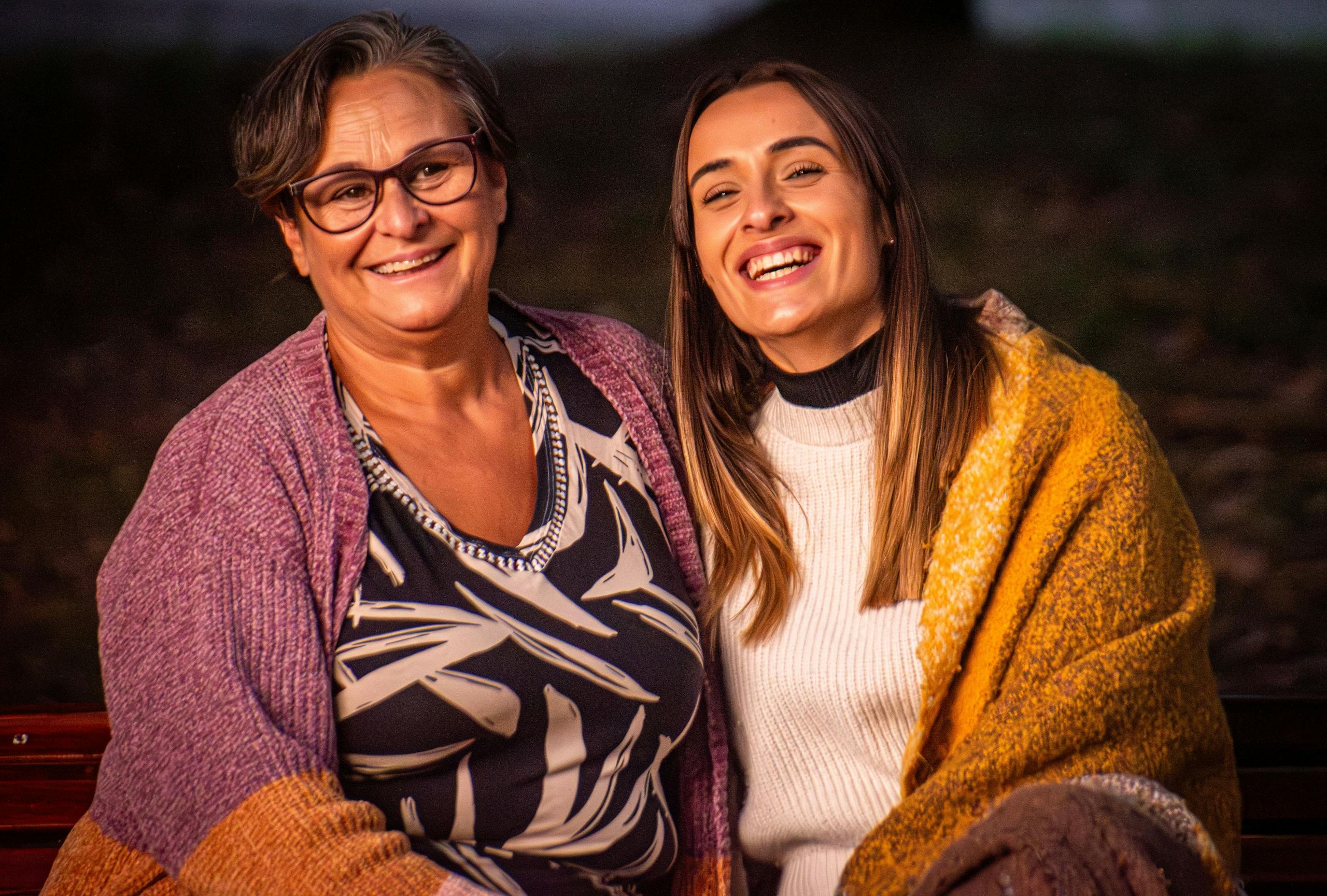 Smiling mother and daughter sitting outdoors wrapped in cozy blankets.