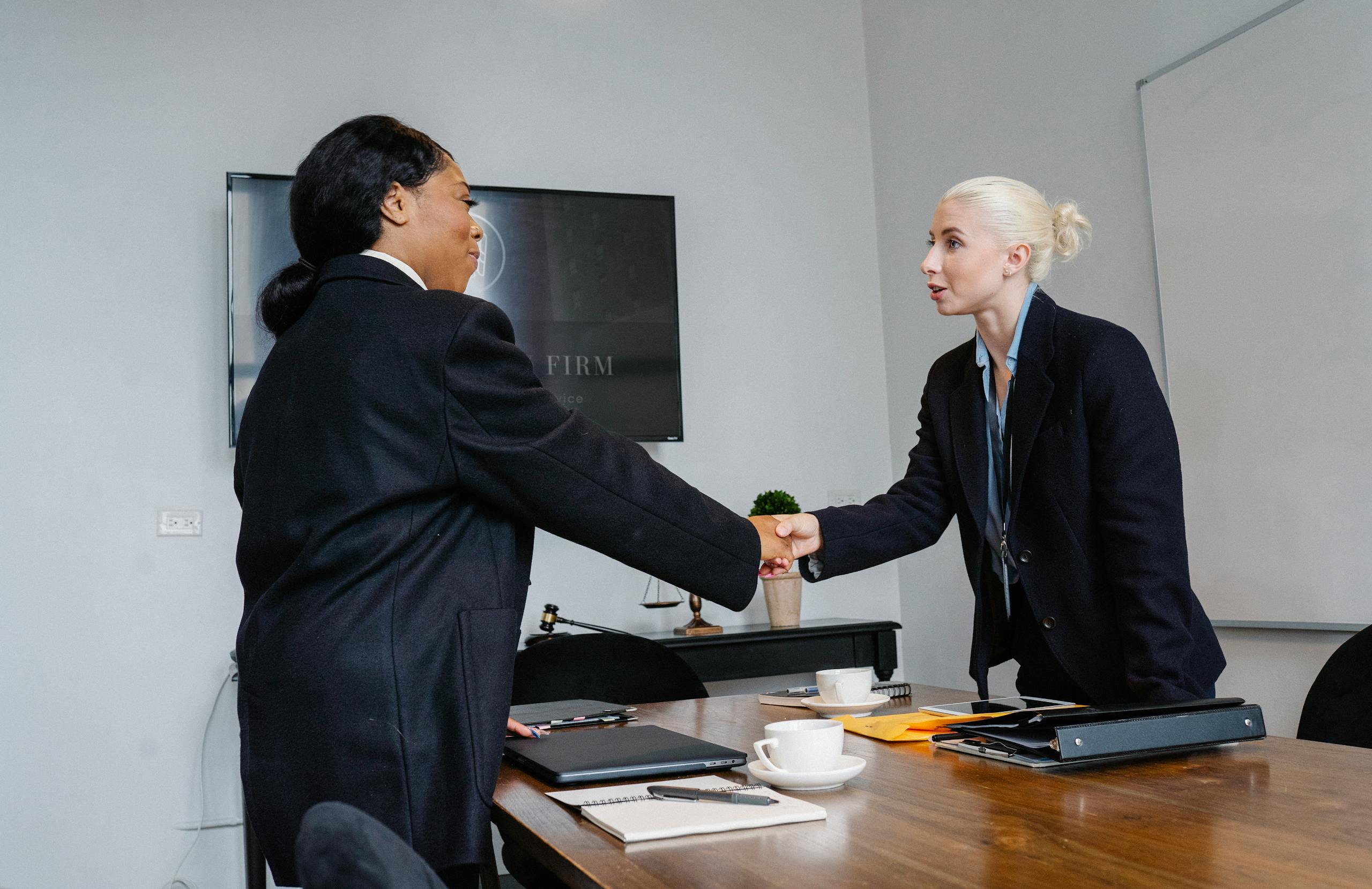 Smiling multiracial women in formal wear standing near table with documents and shaking hands after successful deal
