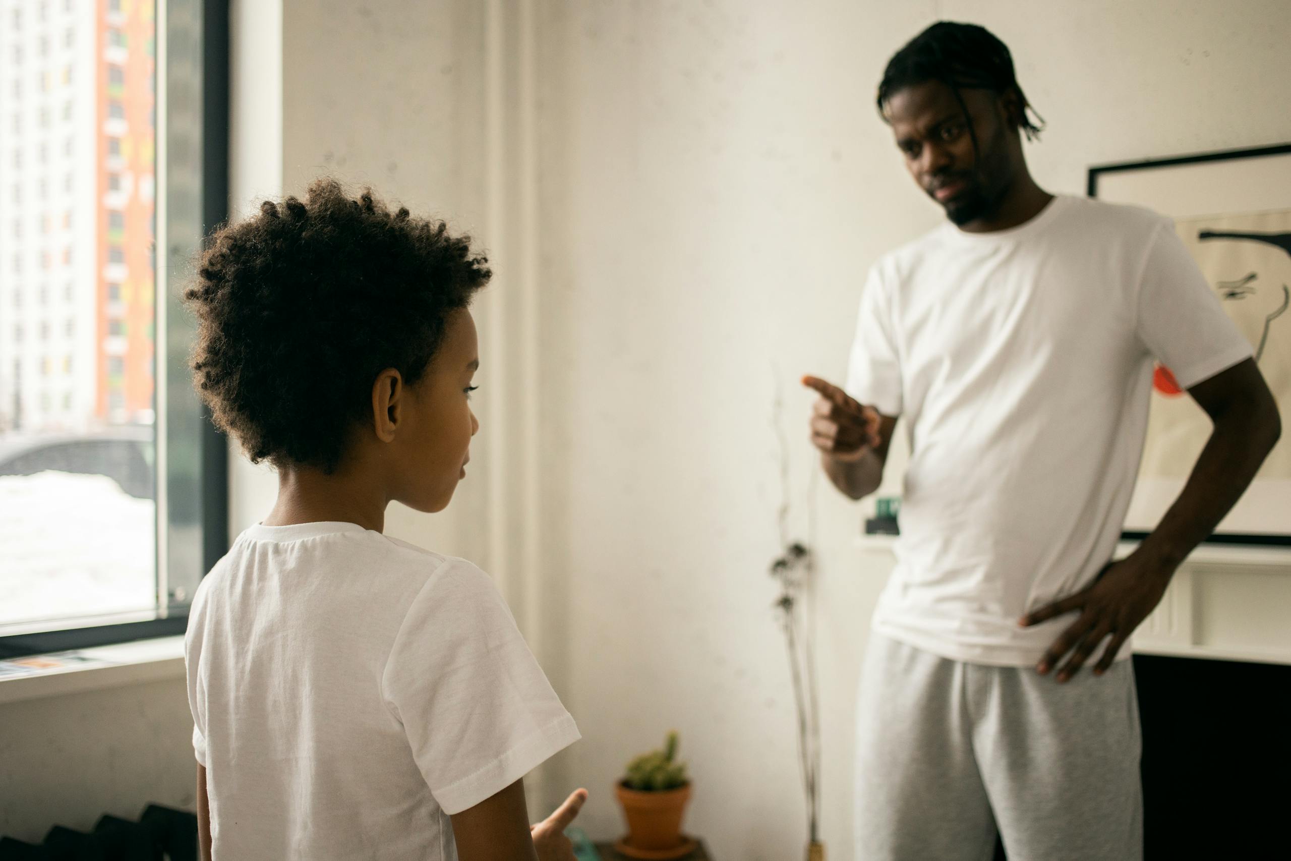 Strict young African American father in white t shirt pointing finger and scolding upset little son at home