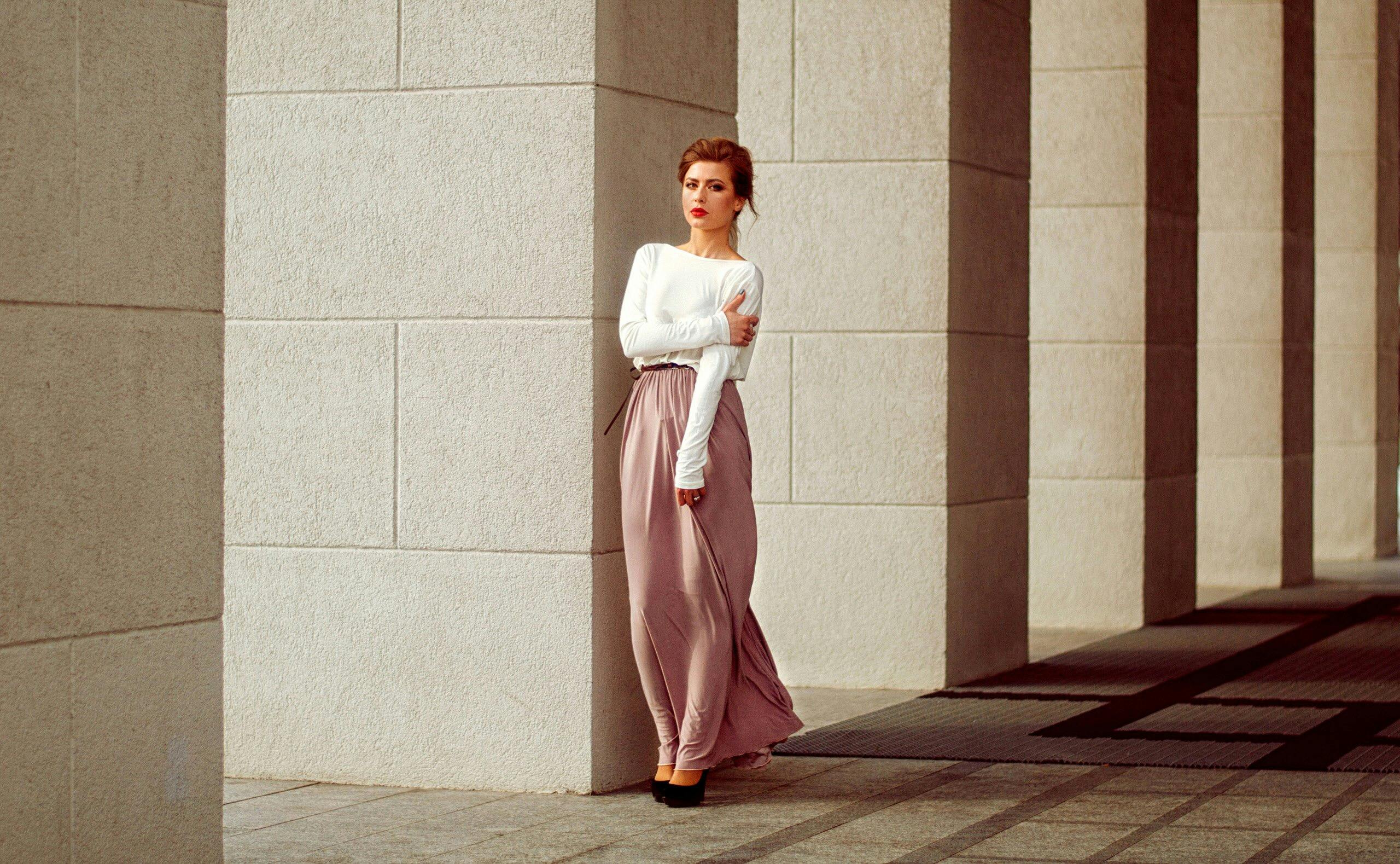 Stylish woman in pink skirt leans against a stone pillar in sunlight.