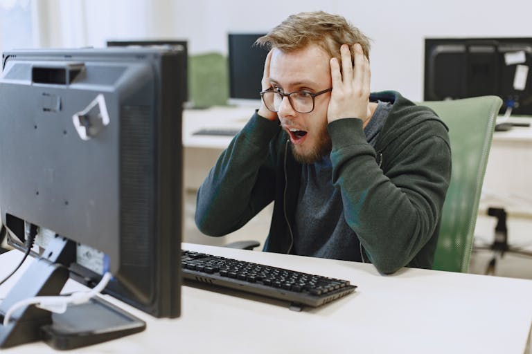 Surprised man in hoodie reacting to shocking news on computer monitor while sitting in a modern office.