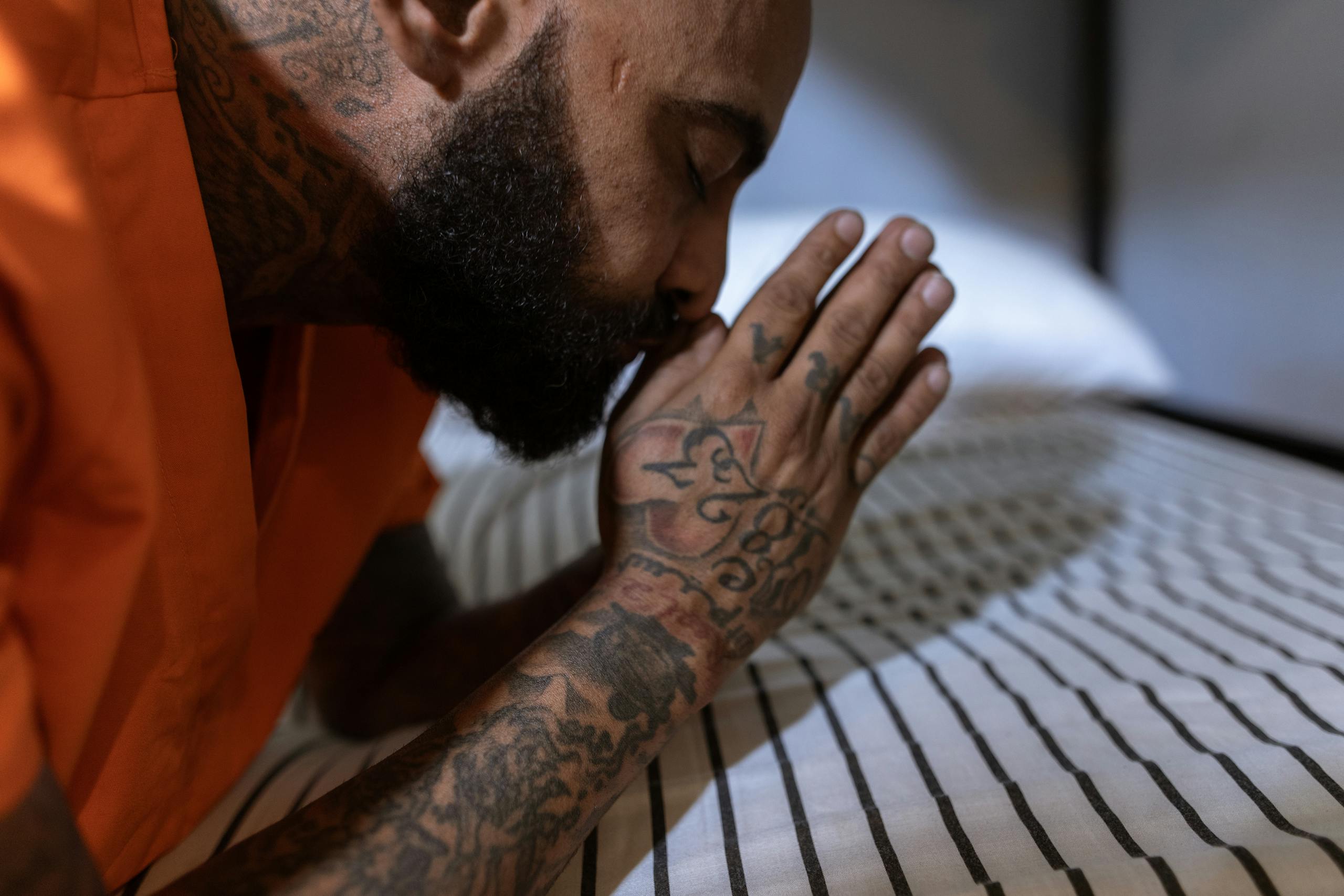 Tattooed inmate in orange uniform kneeling by bed, hands clasped in prayer.