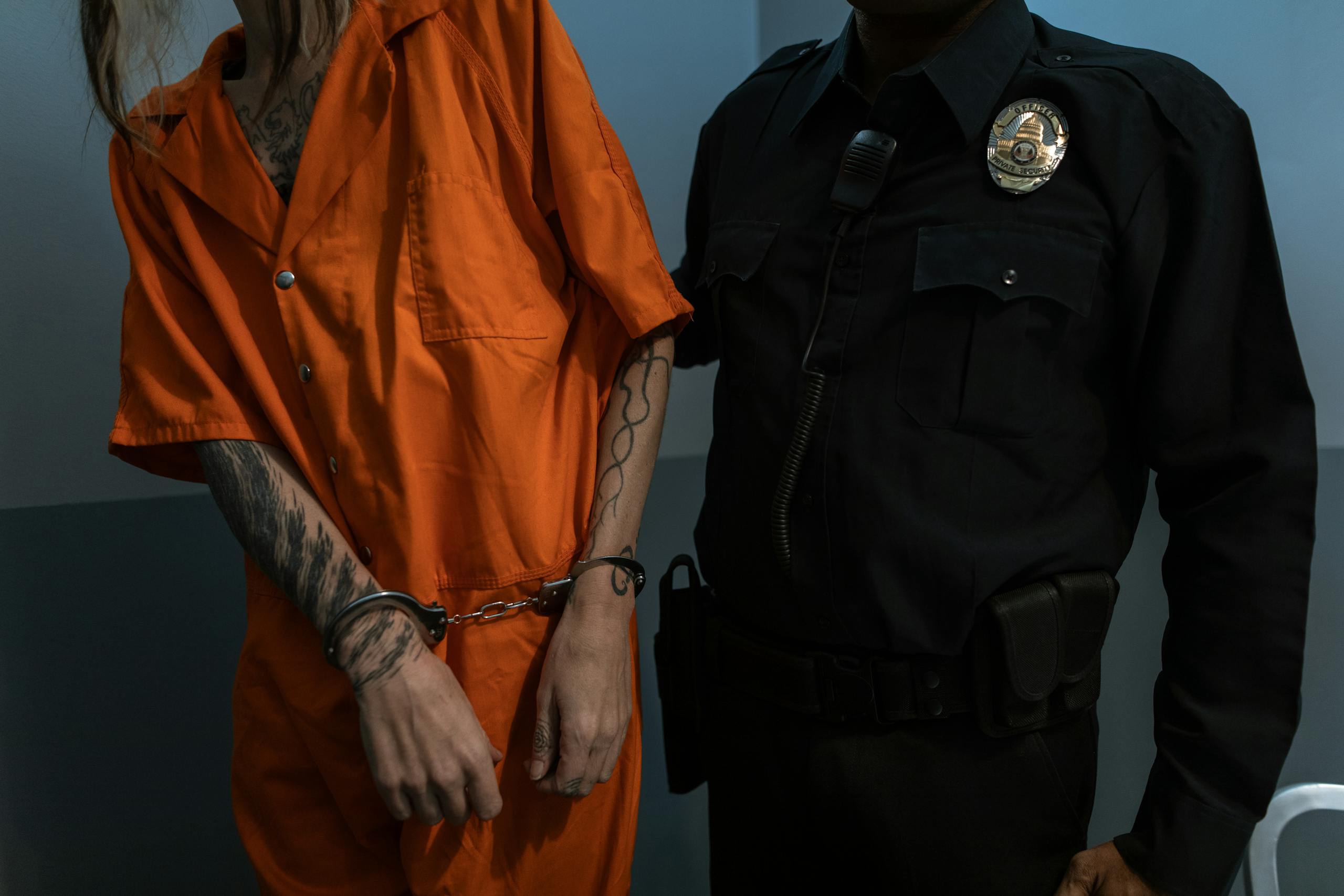 Tattooed prisoner handcuffed in orange jumpsuit beside uniformed officer, highlighting law enforcement.