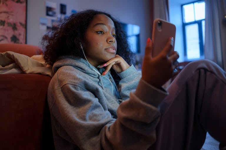 Teenage girl using smartphone indoors with earphones, wearing a hoodie, and looking relaxed.