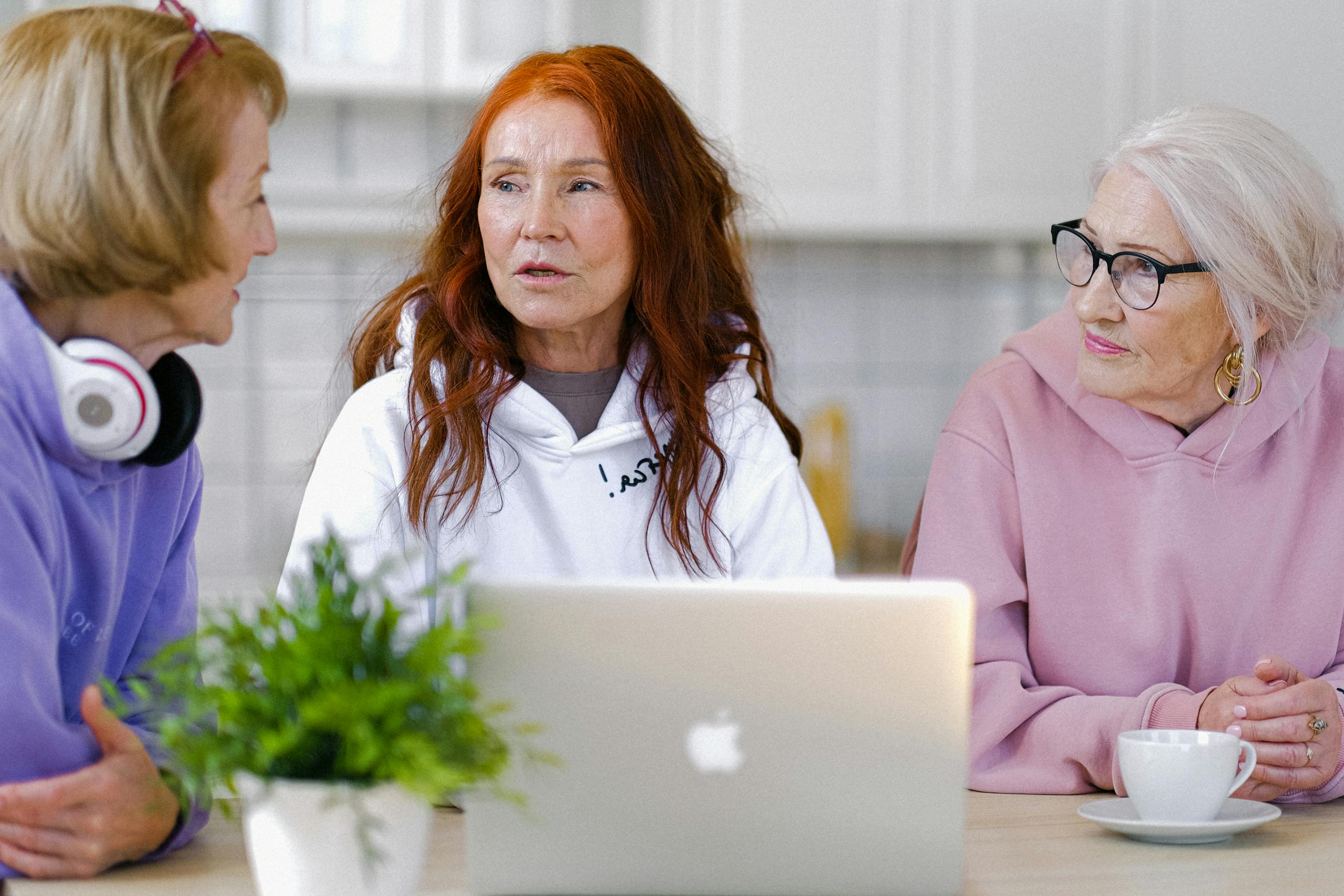 Thoughtful senior women in colorful hoodies sitting at table with cups of tea and opened netbook and communicating in modern apartment with light interior