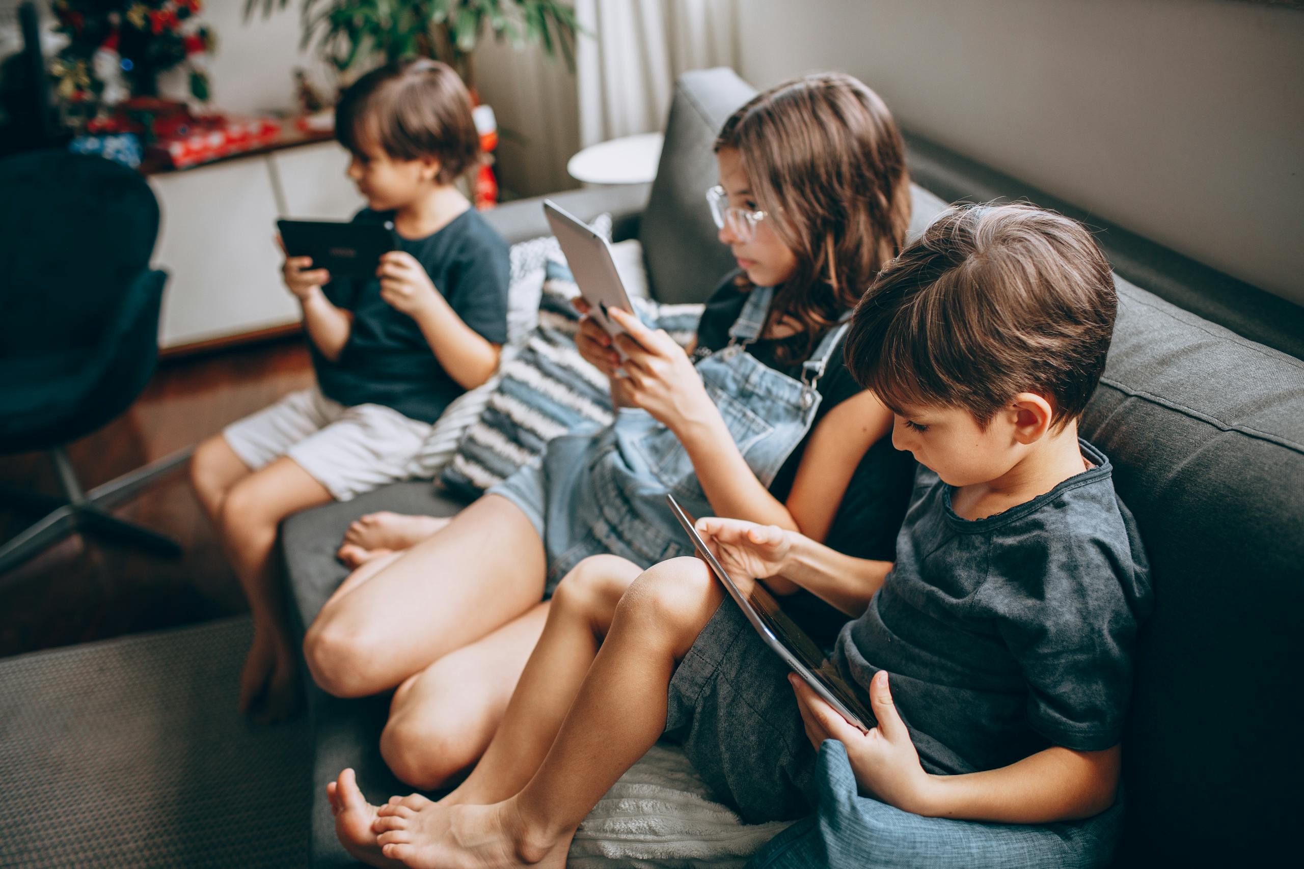 Three children using digital tablets while sitting together on a cozy sofa, enjoying technology indoors.