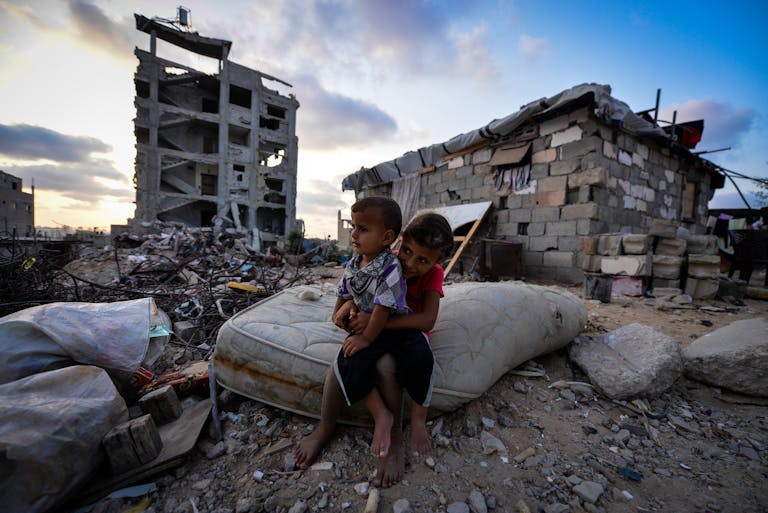 Two children sit on a mattress amidst the ruins of a building in an urban decay setting during early twilight.
