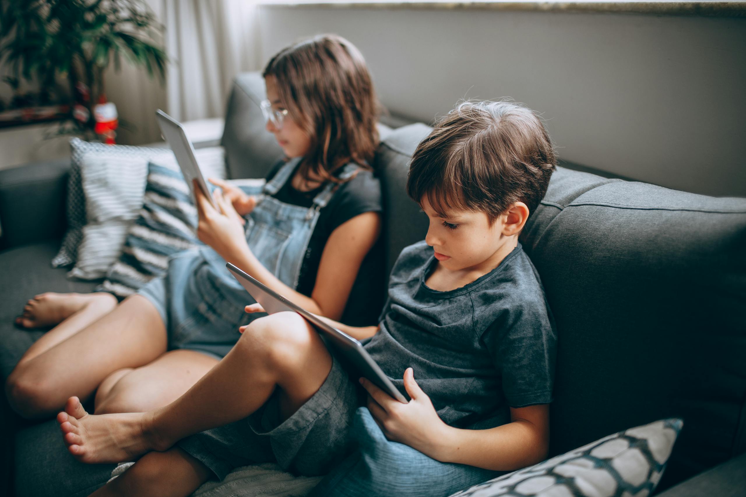 Two children sitting on a sofa using tablets indoors, focused on the screens.