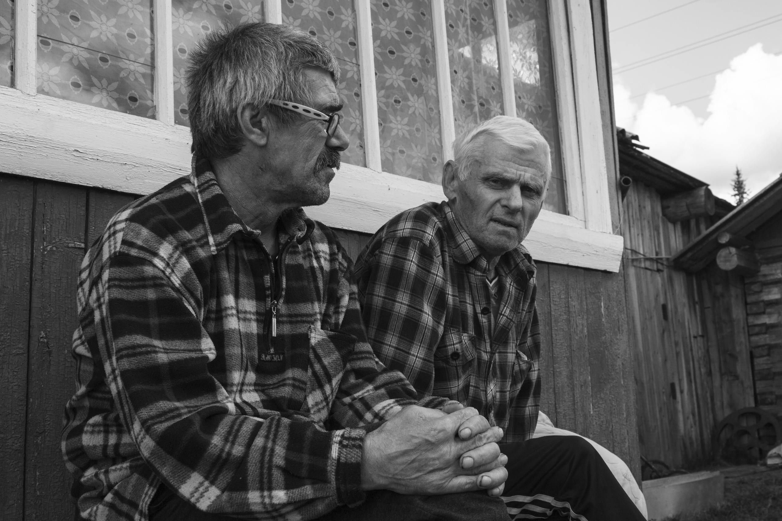 Two elderly men in plaid shirts sitting by a wooden wall in grayscale.