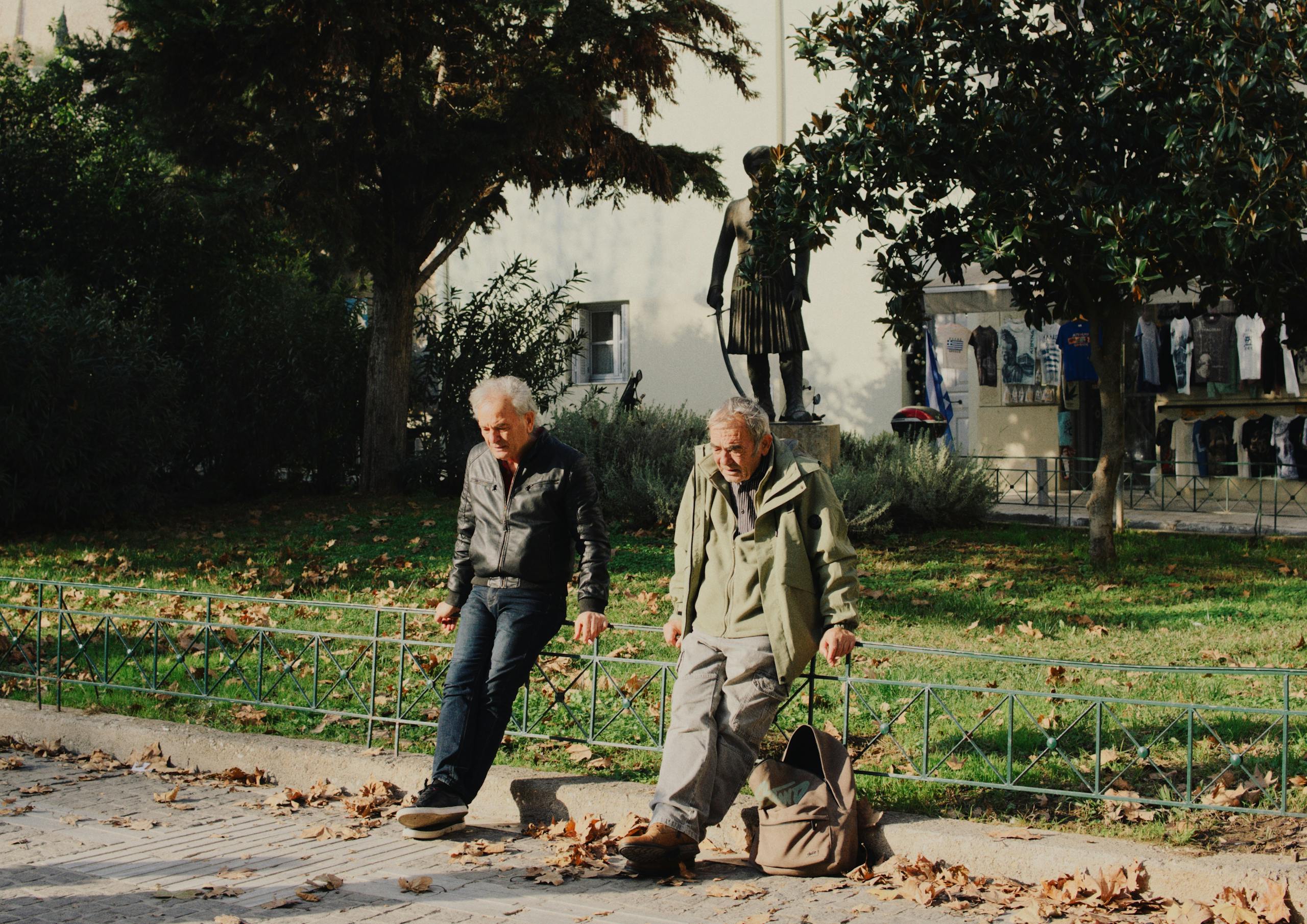 Two elderly men sit in a sunny park with autumn leaves around them, near a statue.