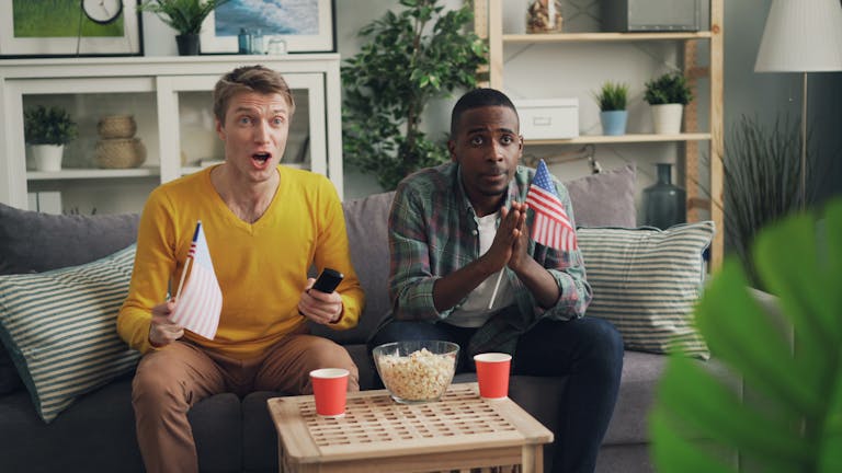 Two excited friends on a couch watching sports, holding USA flags and popcorn.
