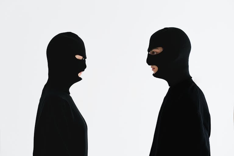 Two individuals in black balaclavas facing each other against a white background.