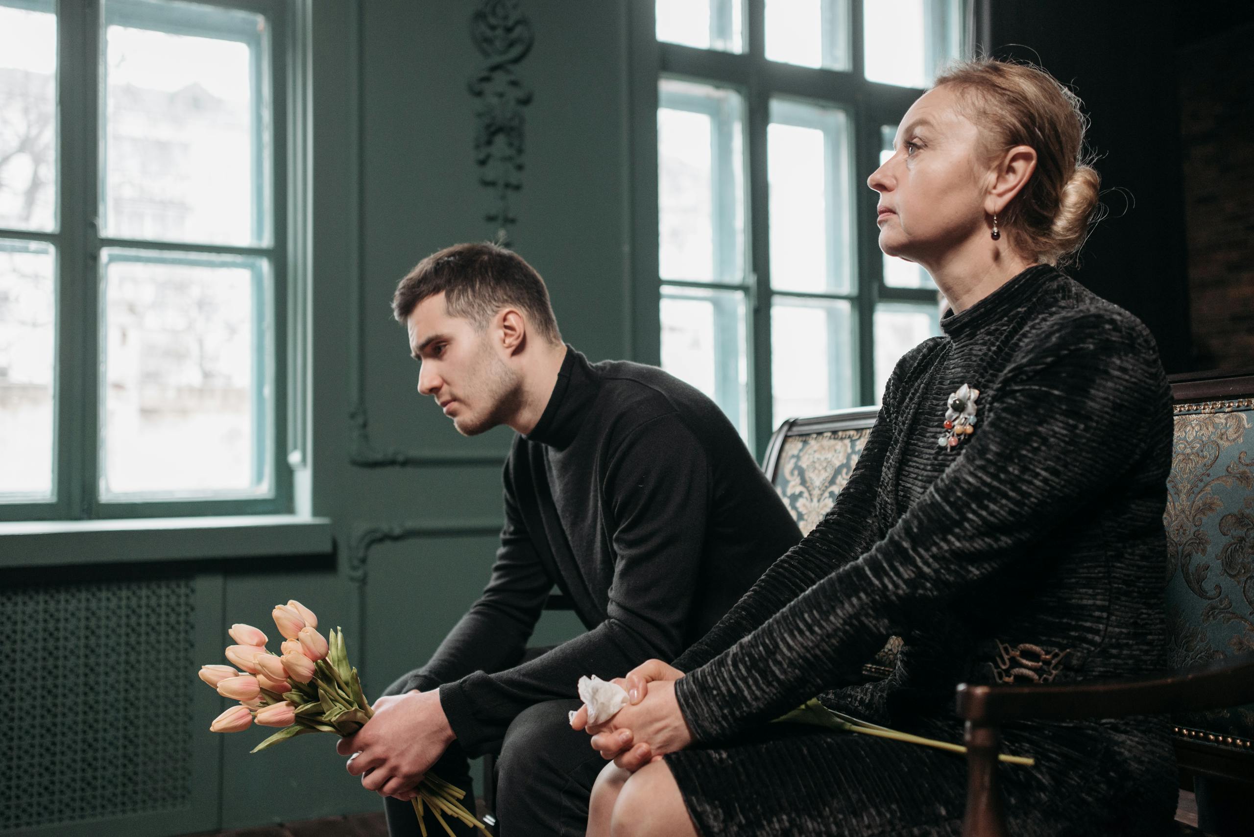 Two individuals in mourning attire sitting solemnly indoors, reflecting sorrow.