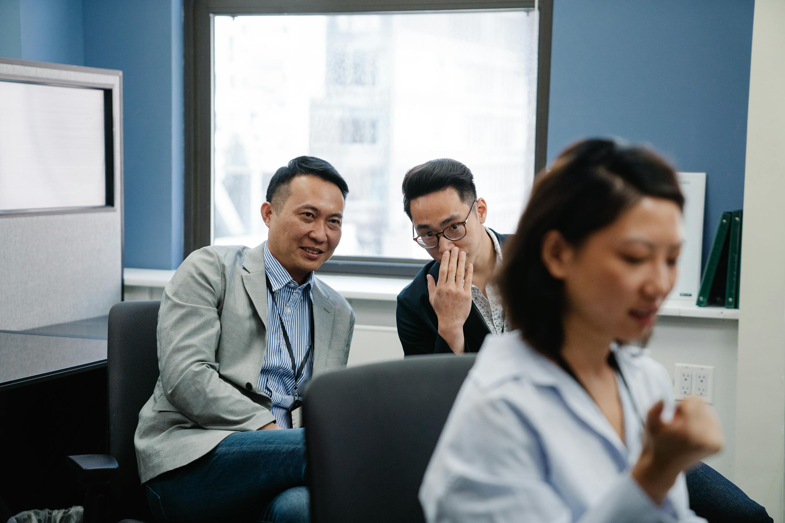 Two male colleagues sharing a secret in an office while a female coworker works nearby.