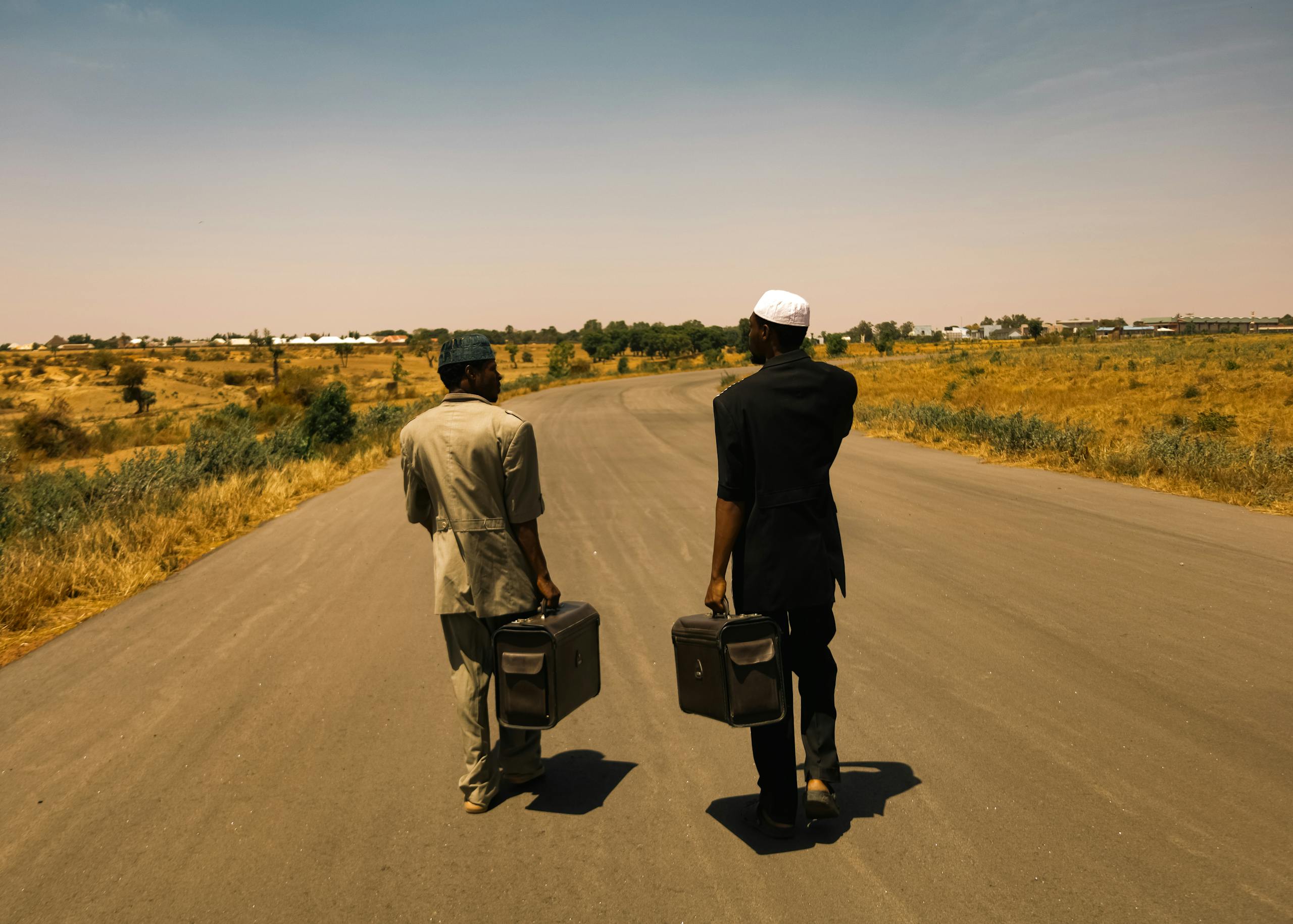 Two men walking on a rural road carrying briefcases on a sunny day, representing journey and companionship.