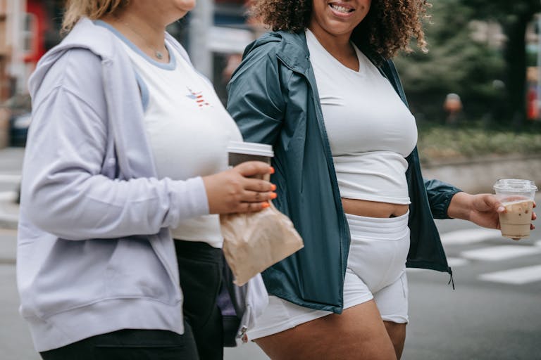 Two women enjoying a walk while holding coffee cups, wearing casual sports attire in a city setting.