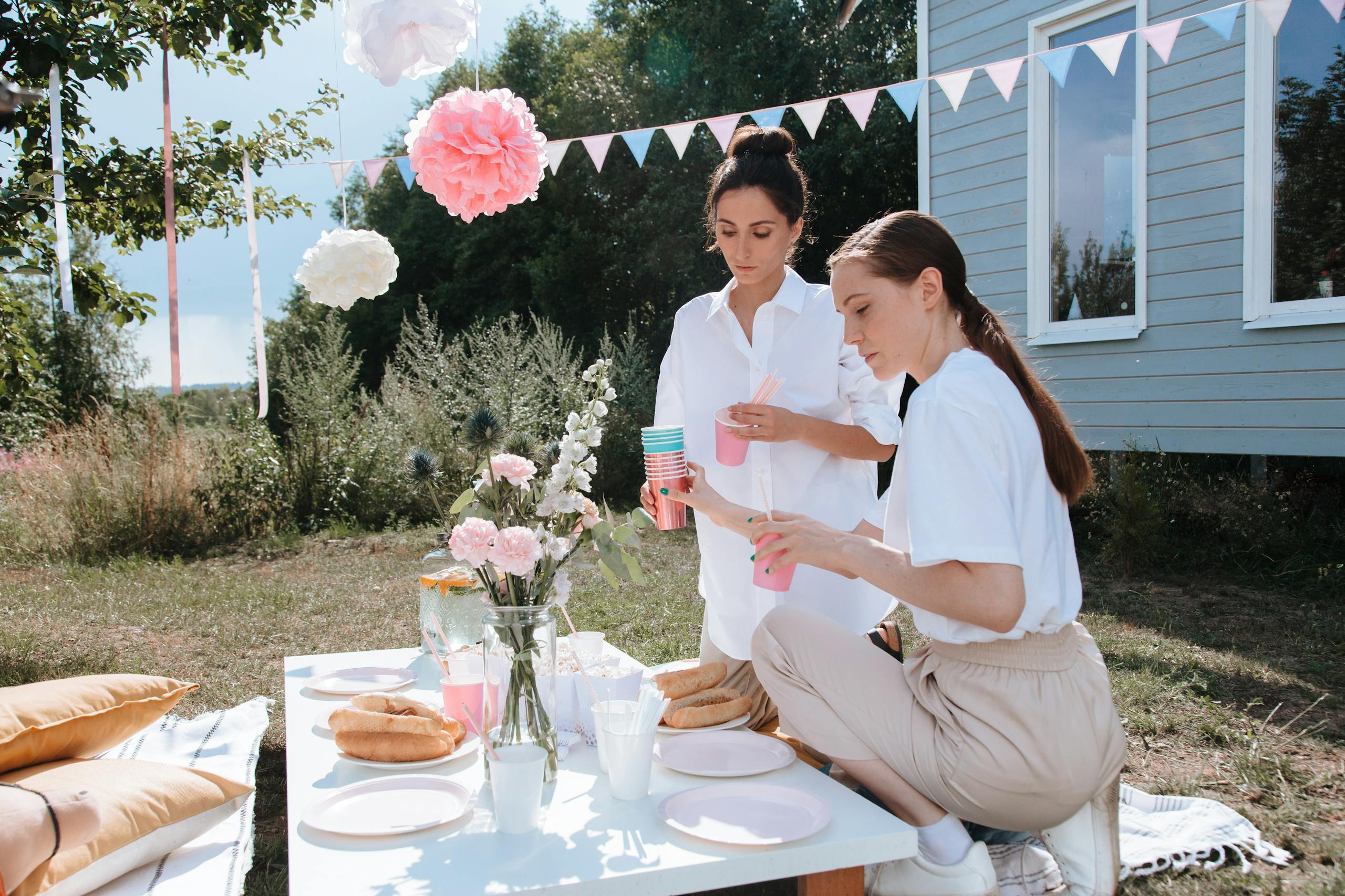 Two women preparing a festive outdoor table setting with breads, drinks, and floral decor.