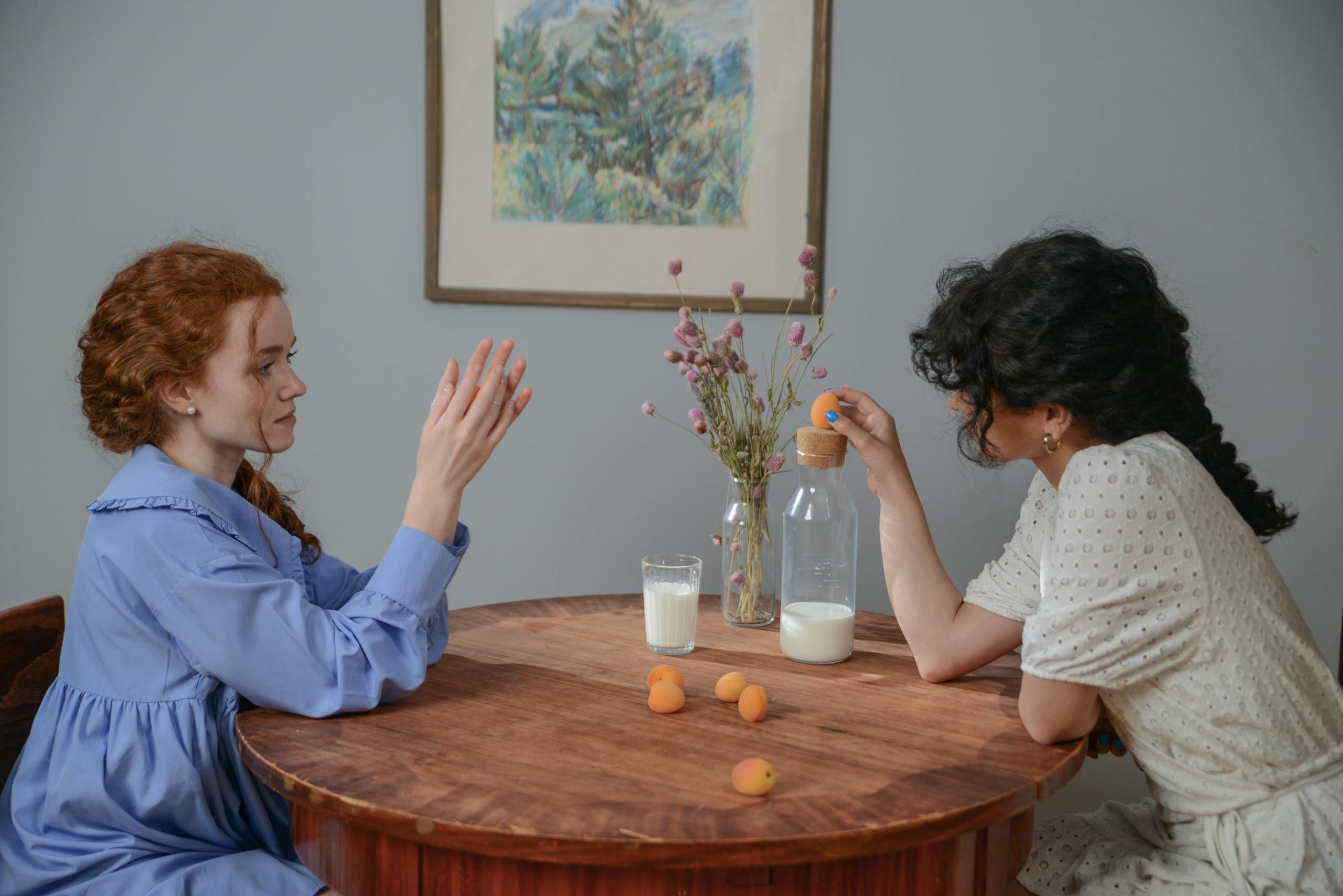 Two women sit at a wooden table with milk and apricots, sharing a calm conversation.